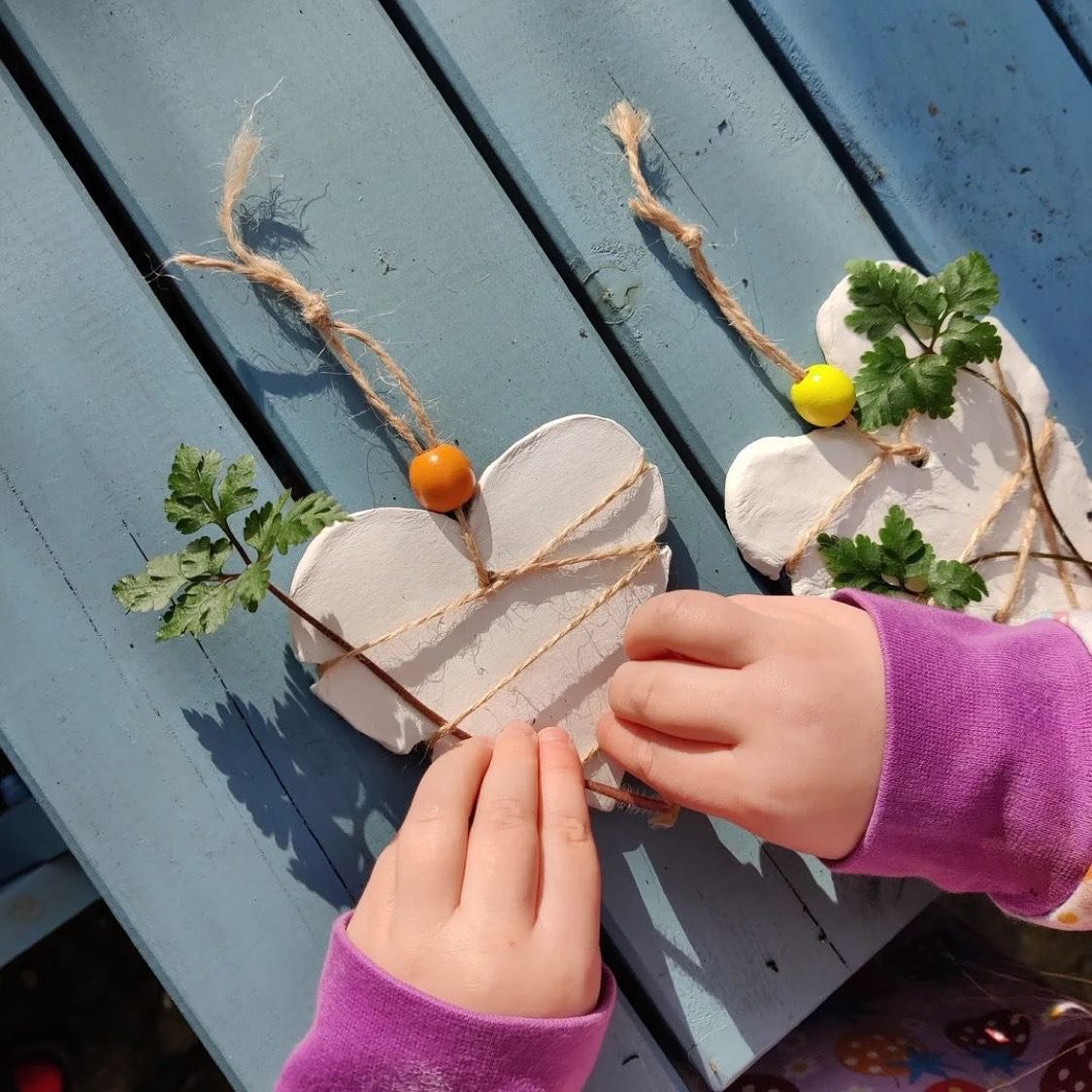 Child's hands attaching leaves and colorful beads to decorative white cutouts, resembling hearts, with twine, on a blue wooden surface.