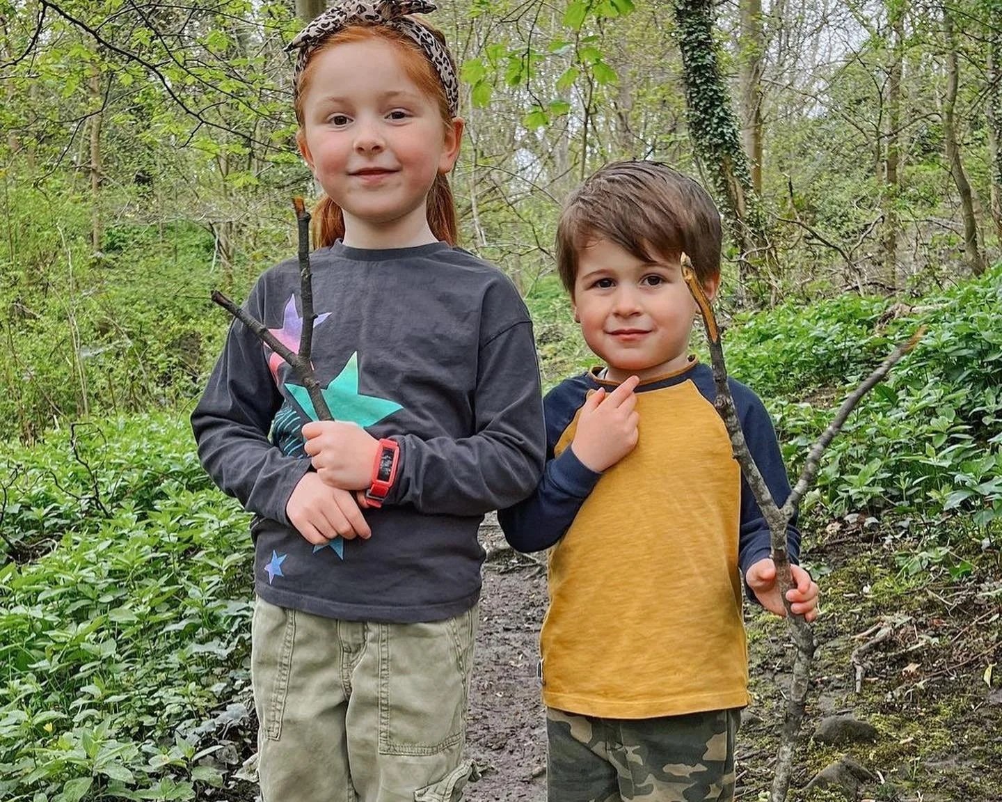 Two young children, a girl with red hair and a boy with brown hair, standing on a wooded trail during daytime, each holding a small tree branch. The girl is wearing a black long sleeve shirt with colorful stars, khaki pants, and a leopard print headband. The boy is wearing a yellow and navy long sleeve shirt and camo pants, with one hand touching his chin. They are surrounded by green foliage and trees.