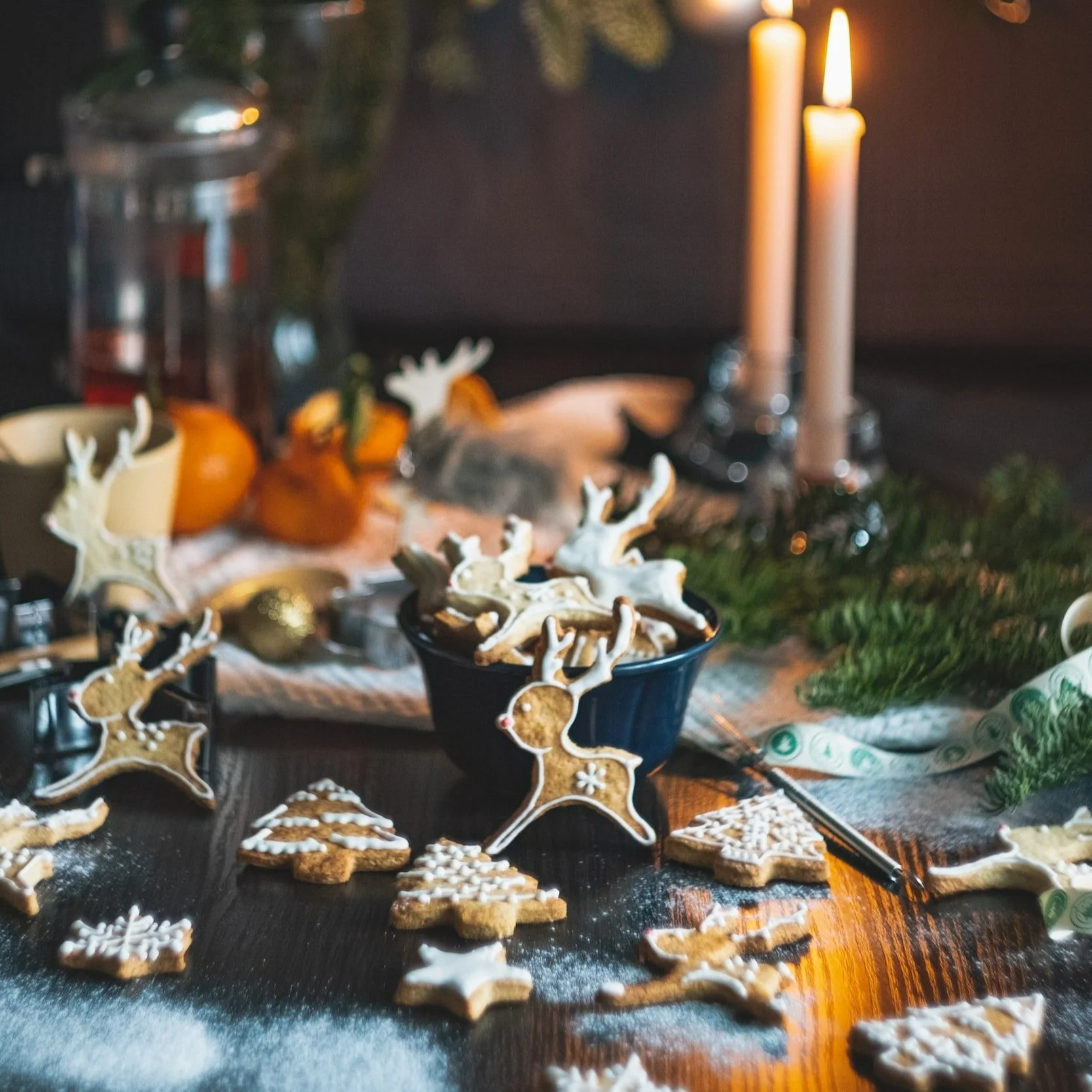 Gingerbread reindeer, Christmas trees, and stars scattered on a table and dusted with icing sugar. Lit taper candles, satsumas and foliage in the background .
