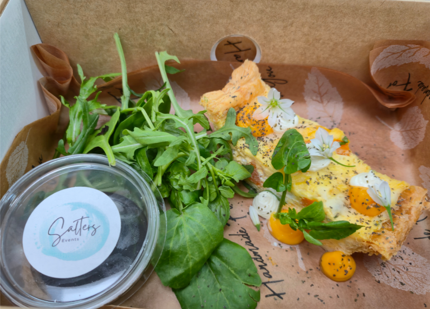 Rich butternut squash and goats cheese tart presented in eco platter box with bright green salad leaves and vinaigrette in a branded pot