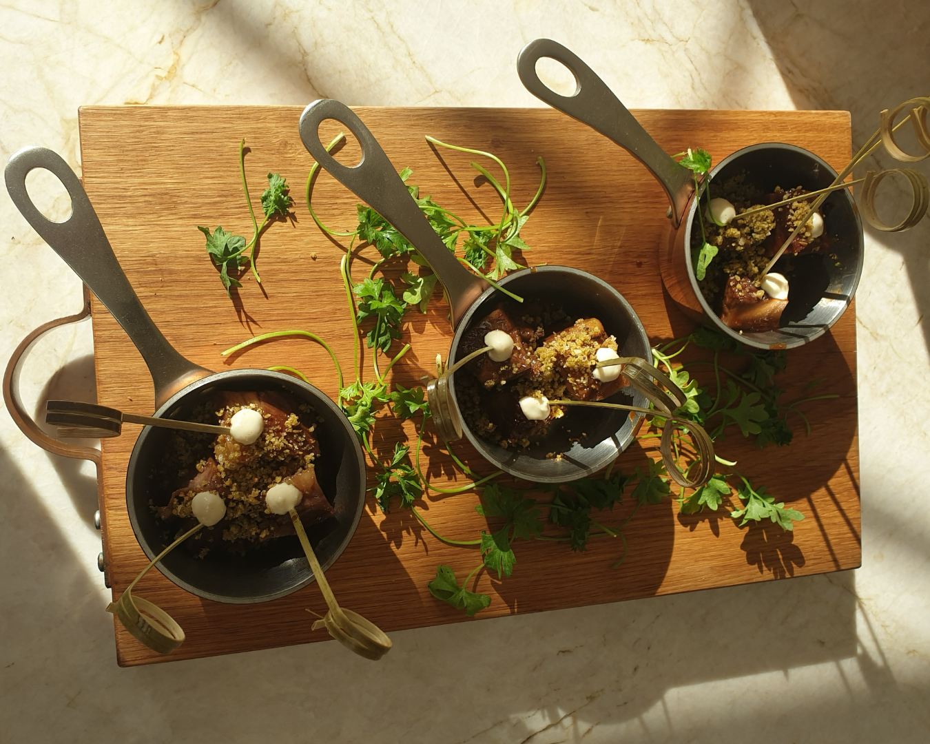 Small copper and silver pans filled with sticky BBQ beef rib canapes, topped with horseradish crème and parsley presented on a wooden board, sitting on a marble counter top.