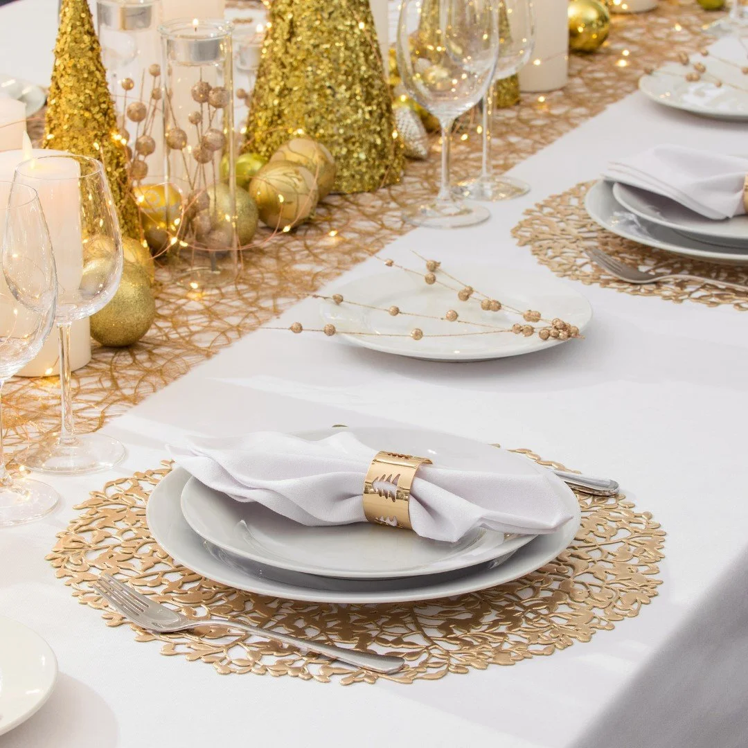 Christmas table laid with a white tablecloth, napkins in gold napkin holders, and  white plates. Lots of gold Christmas decorations, small fairy lights, and glasses.
