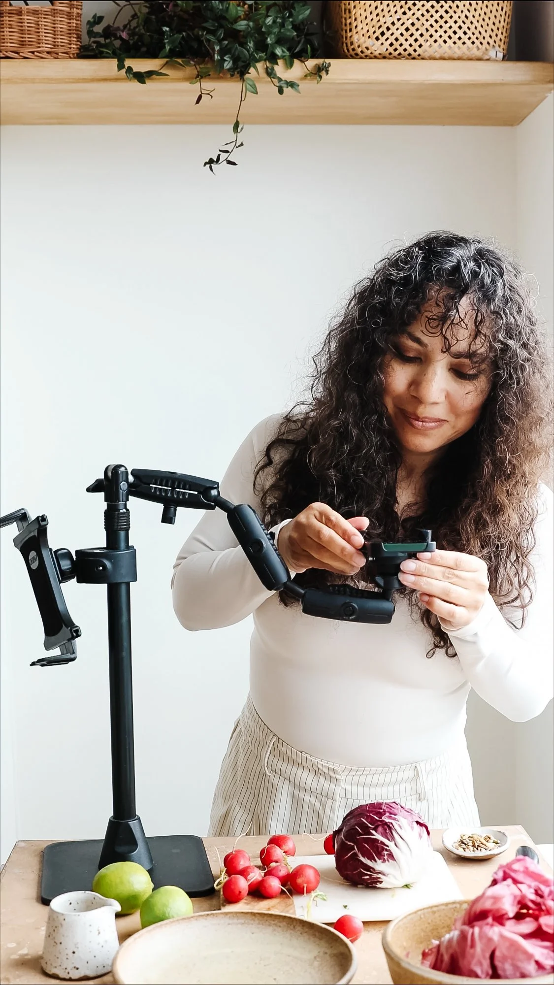 Blog post image showing author Kimberly adjust her phone on a stand aiming down towards a table strewn with ingredients for a recipe reel shoot