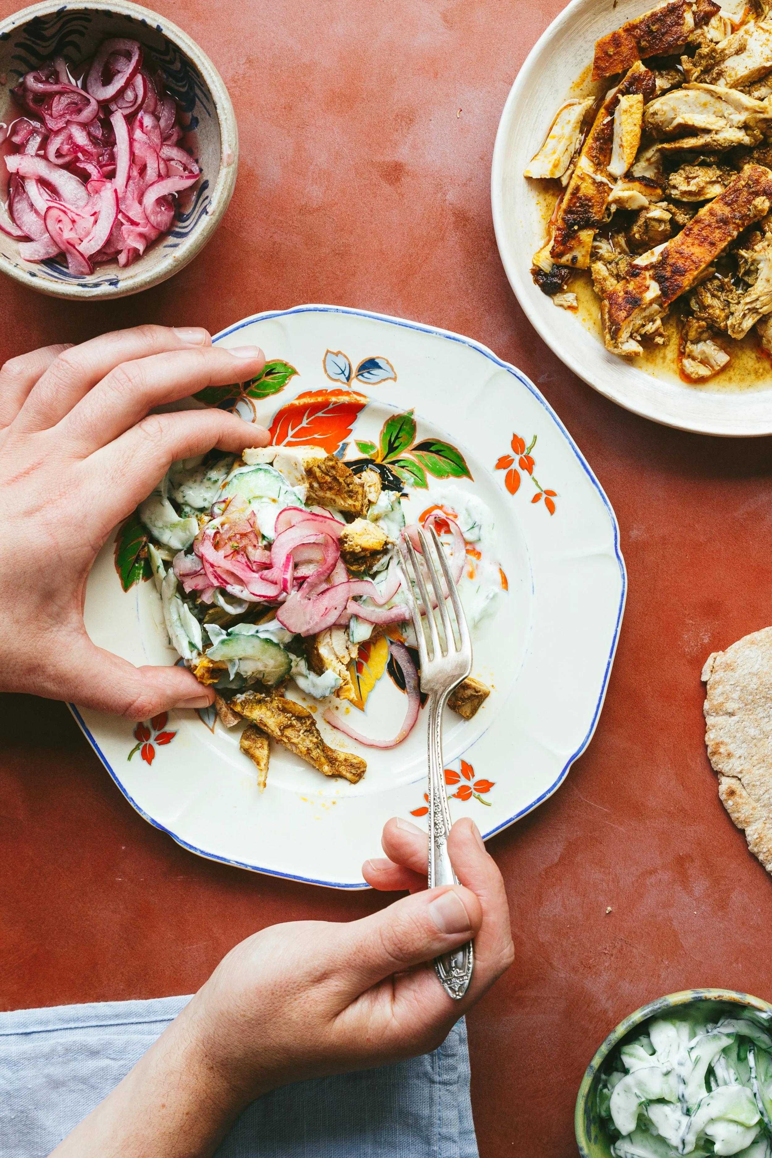 Kimberly Espinel portfolio image showing one hand holding a piece of pitta bread loaded with grilled strips of something meaty, tzatziki and pickled onions, while the other hand is holding a fork.