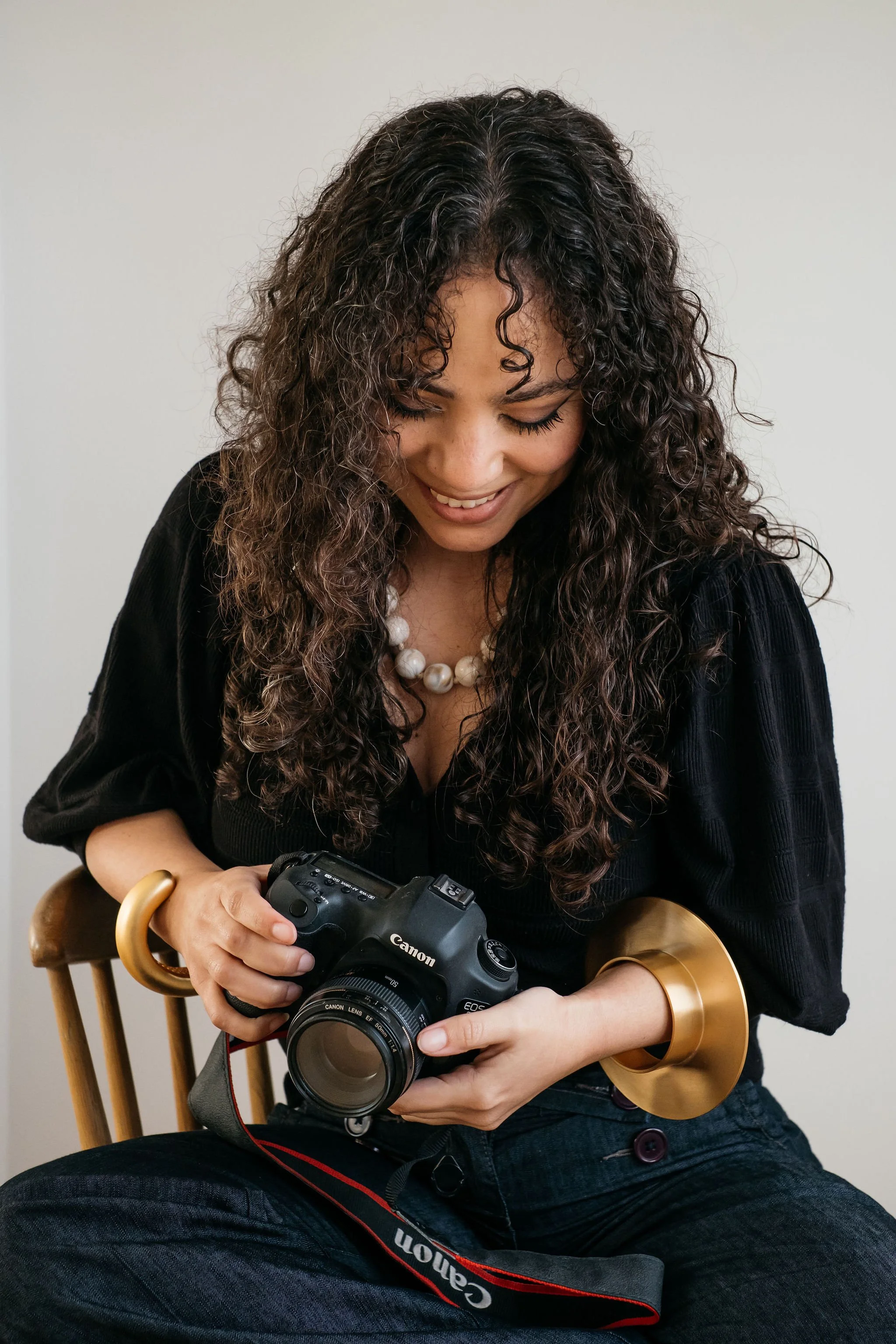 Profile image of Kimberly Espinel sitting in a chair looking down at the camera in her hands and smiling.