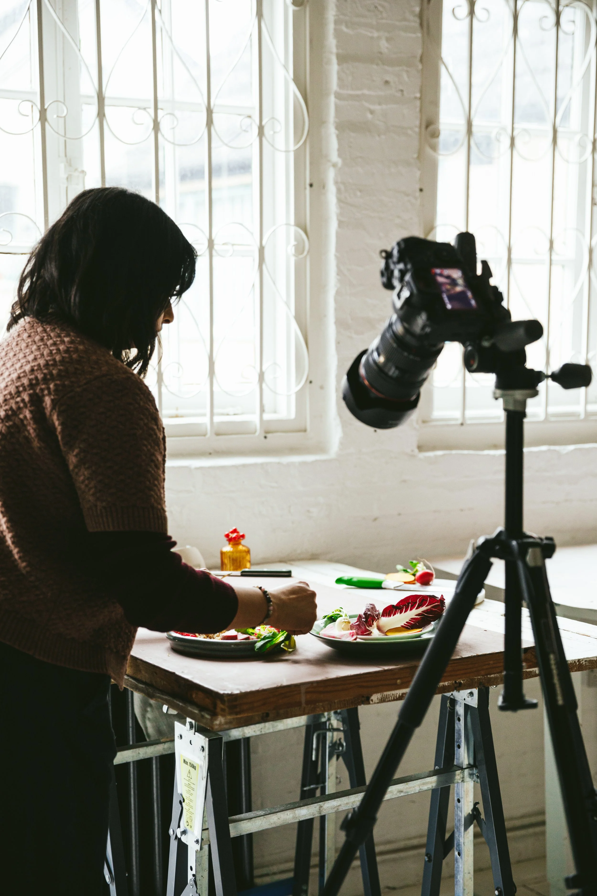 Blog post image showing a person styling a food scene, with a camera on a tripod set up at their side.
