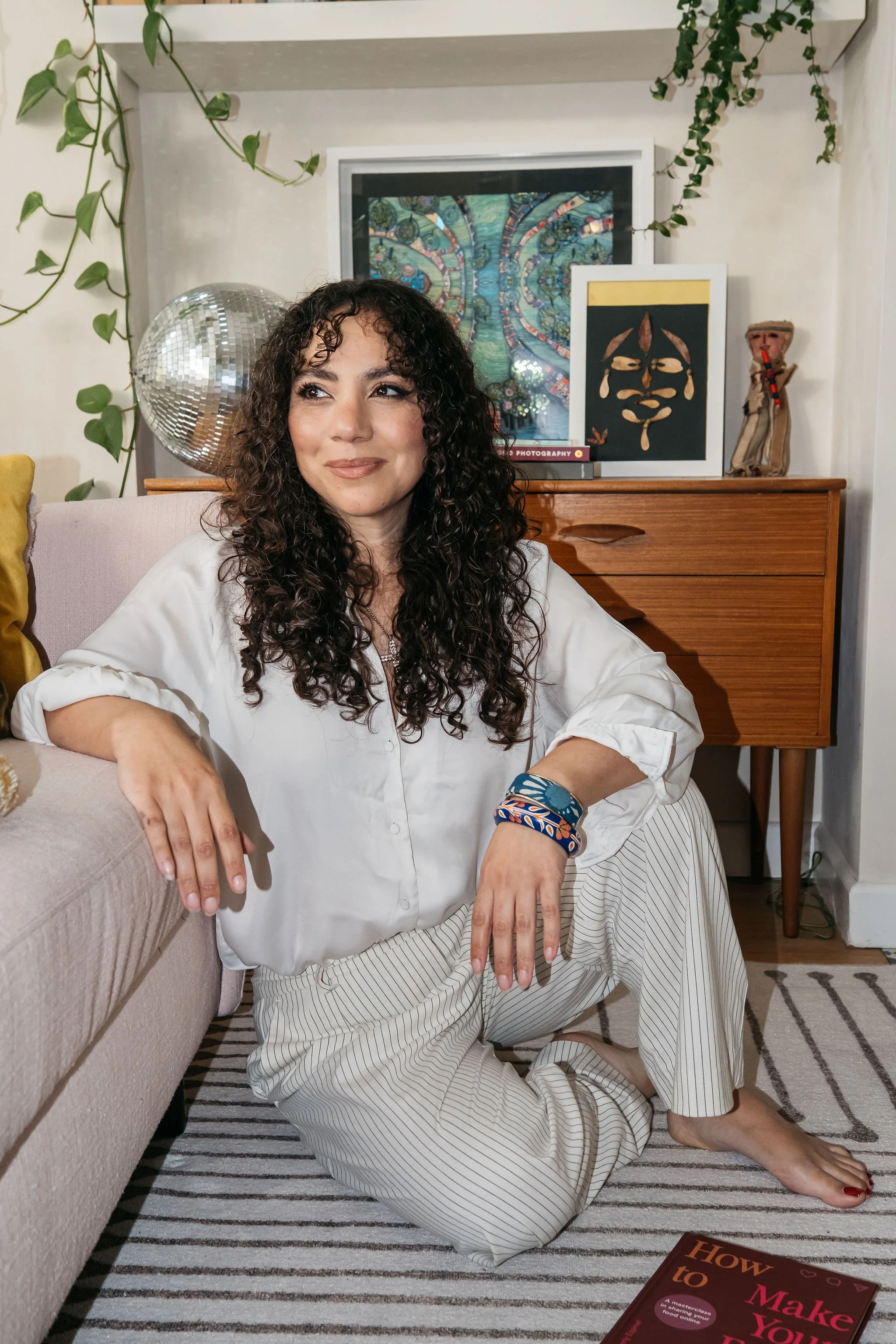 Profile image of food photographer, business mentor and author, Kimberly Espinel, sitting on the floor wearing white linen trousers and shirt, hair worn long and curly, gazing to her right and smiling.
