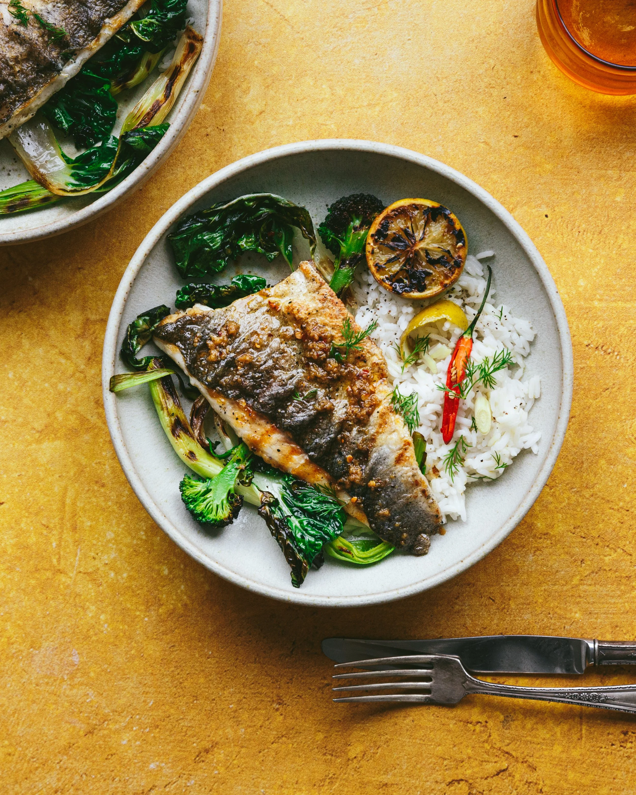 Kimberly Espinel Portfolio image showing a ceramic dish with a serving of grilled fish over rice with charred green vegetables, garnished with charred lemon and a chilli pepper.