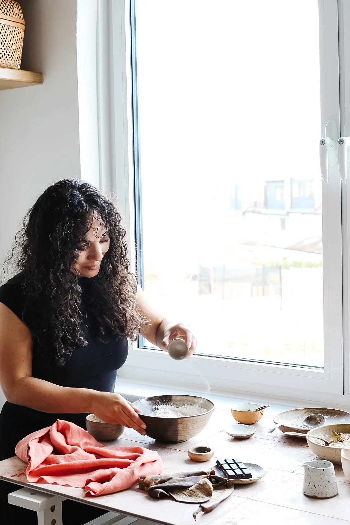 Image shows teacher Kimberly Espinel shaking flour over a bowl as part of a food scene beside a large bright window