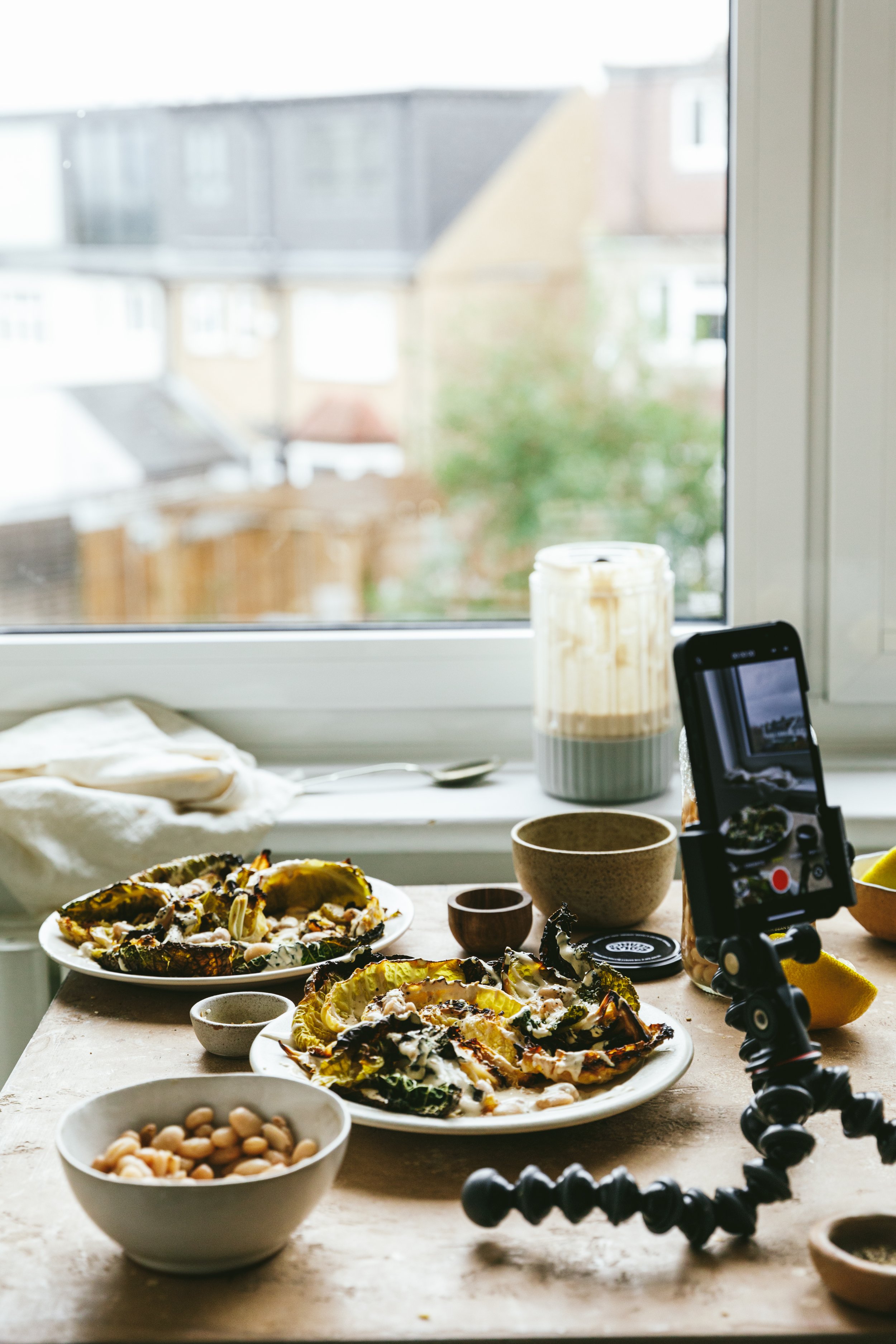 Blog post image showing a camera holder with a phone attached, on a table, filming a food scene with plates of charred cabbage and other ingredients