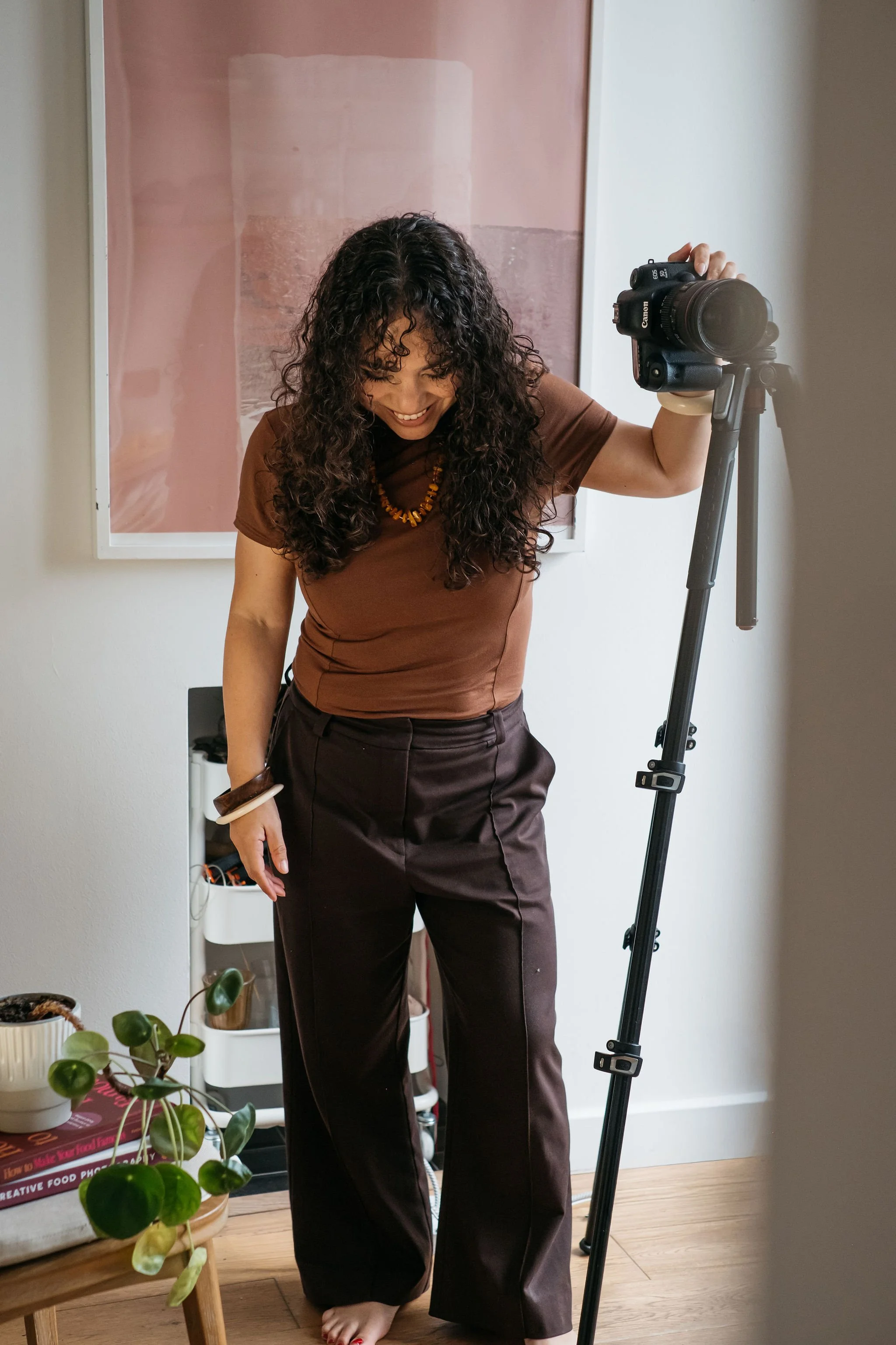 Food photographer and educator Kimberly Espinel standing with her hand resting on her camera mounted on a tipod, while looking down and smiling