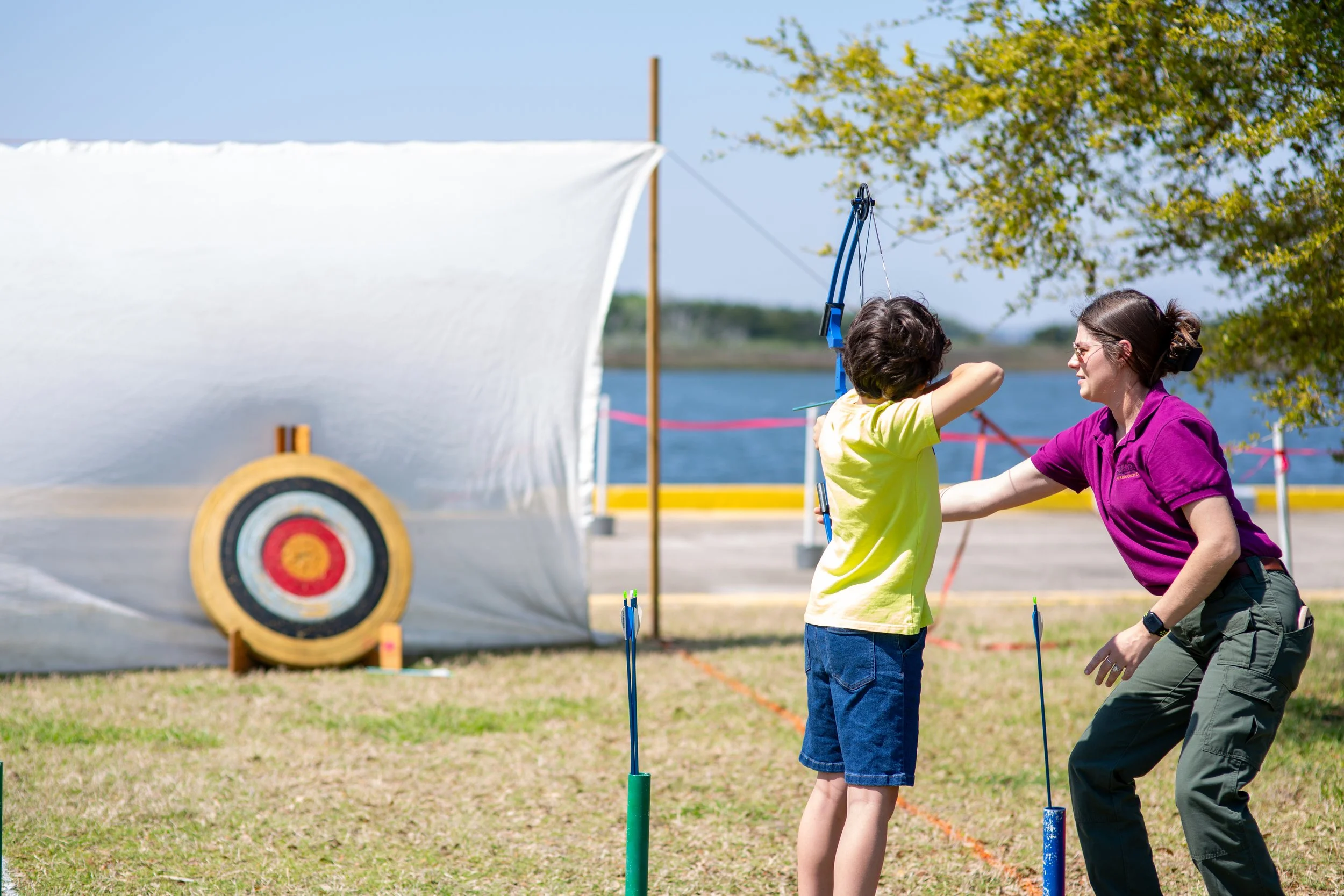 A young boy is shooting a bow and arrow, assisted by a helpful staff member.
