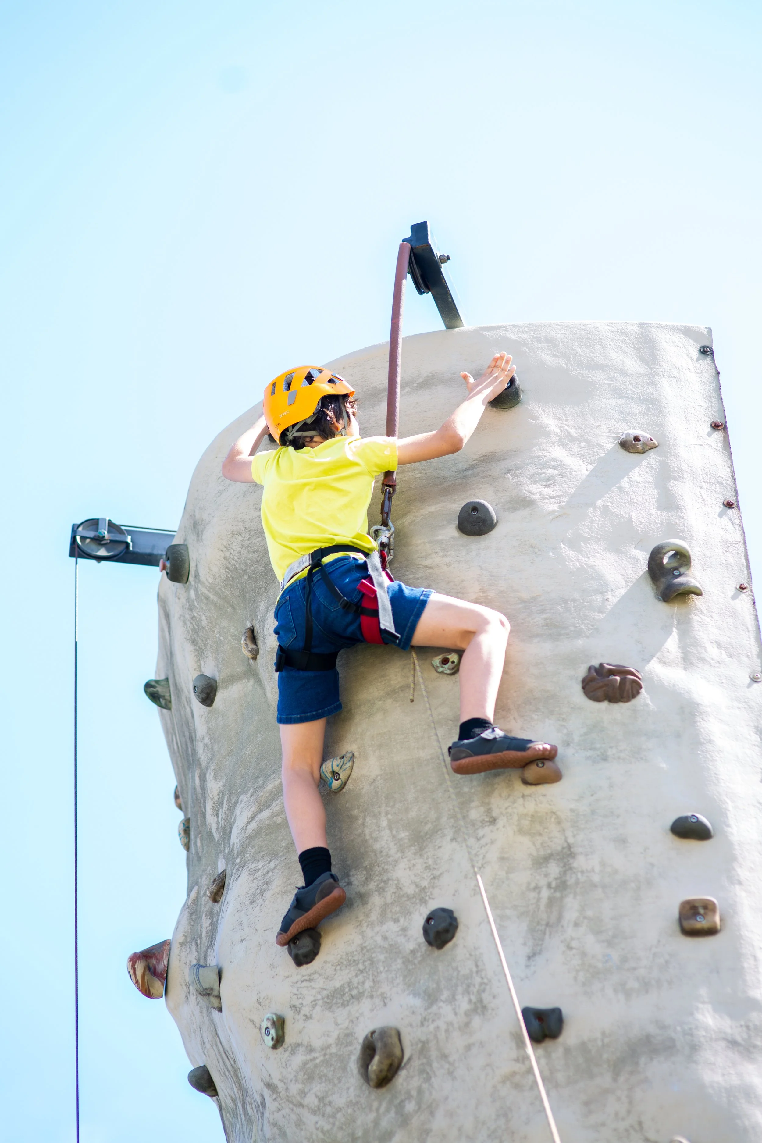 A young boy hits the button at the top of a rock wall at the 2026 Coastfest Even in downtown Brunswick. He is wearing safey equipment.