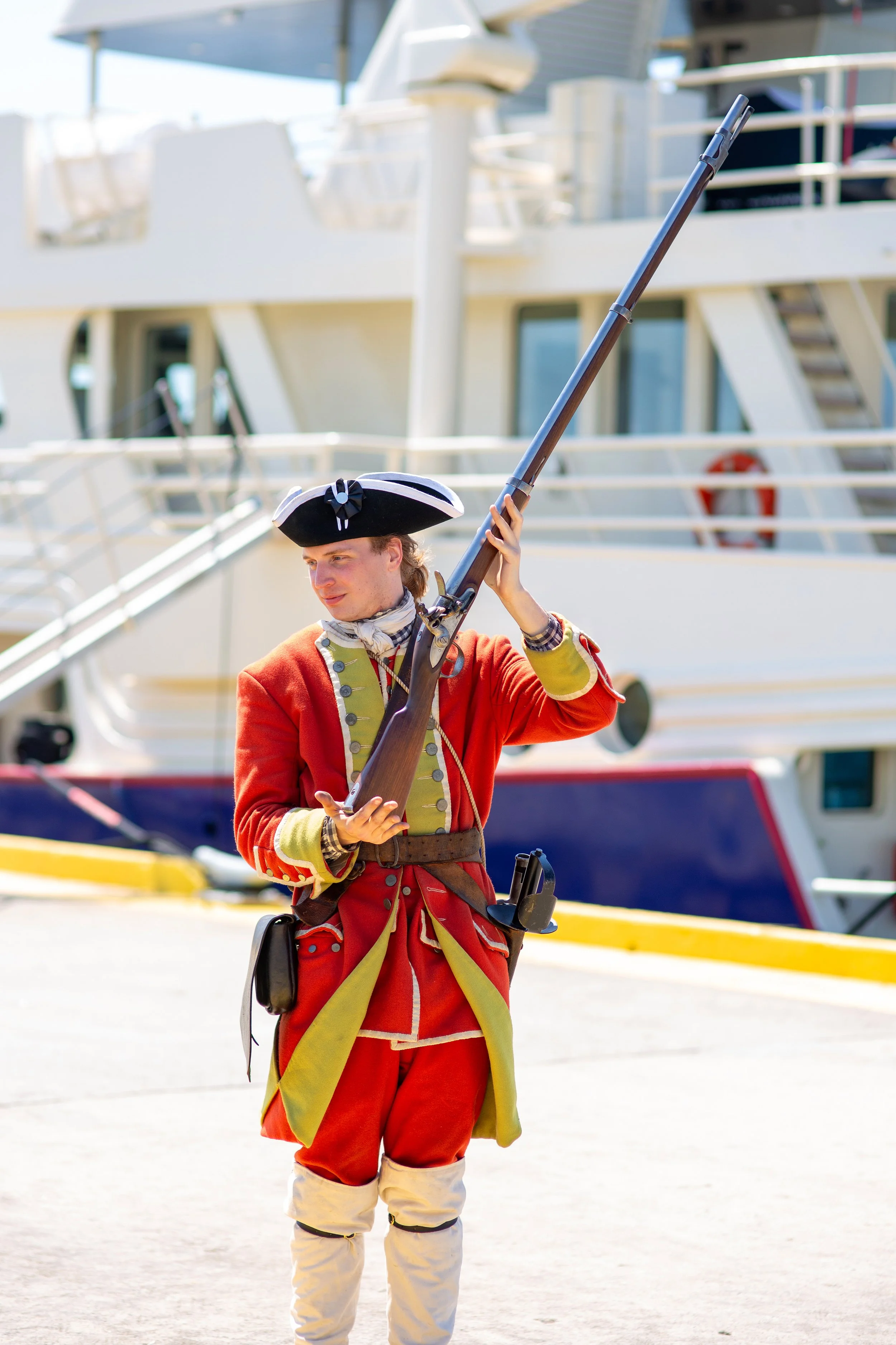 A reinactor stands in front of the Scintilla Maris docked at the Mary Ross Waterfront Park in Brunswick, GA for the 2026 Coastfest event. He is holding a musket and smiling at a young attendee of the event.