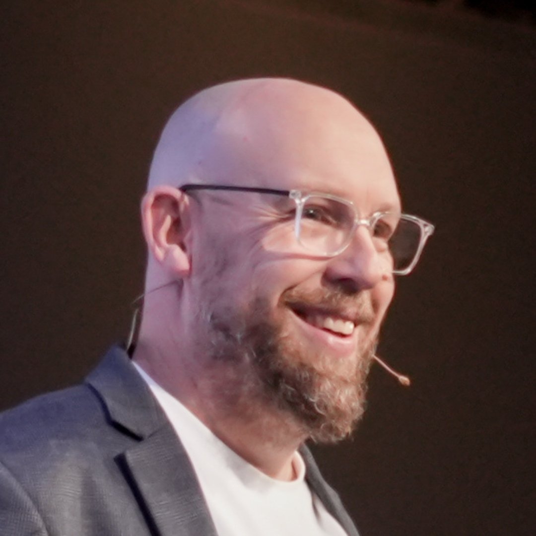 Smiling bald man with glasses and a beard wearing a headset microphone on stage