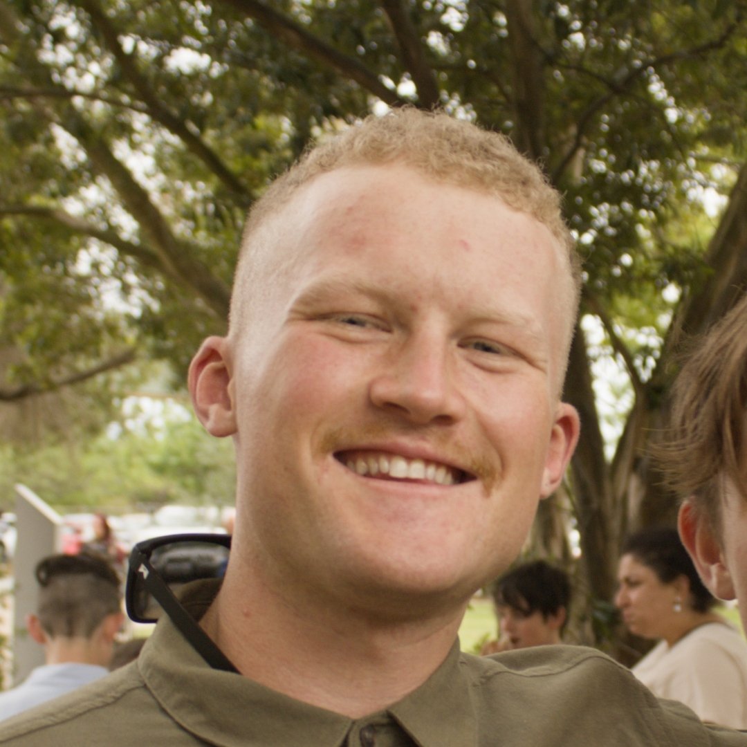 Smiling young man posing outdoors under leafy trees at a social gathering