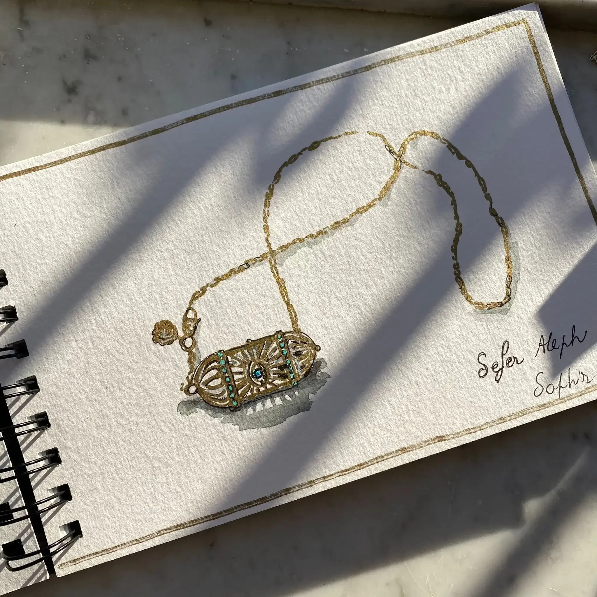 Croquis d'un collier avec une breloque ornée de turquoise, dessiné dans un carnet avec une inscription 'Sefer Aleph Saphir' en bas à droite.