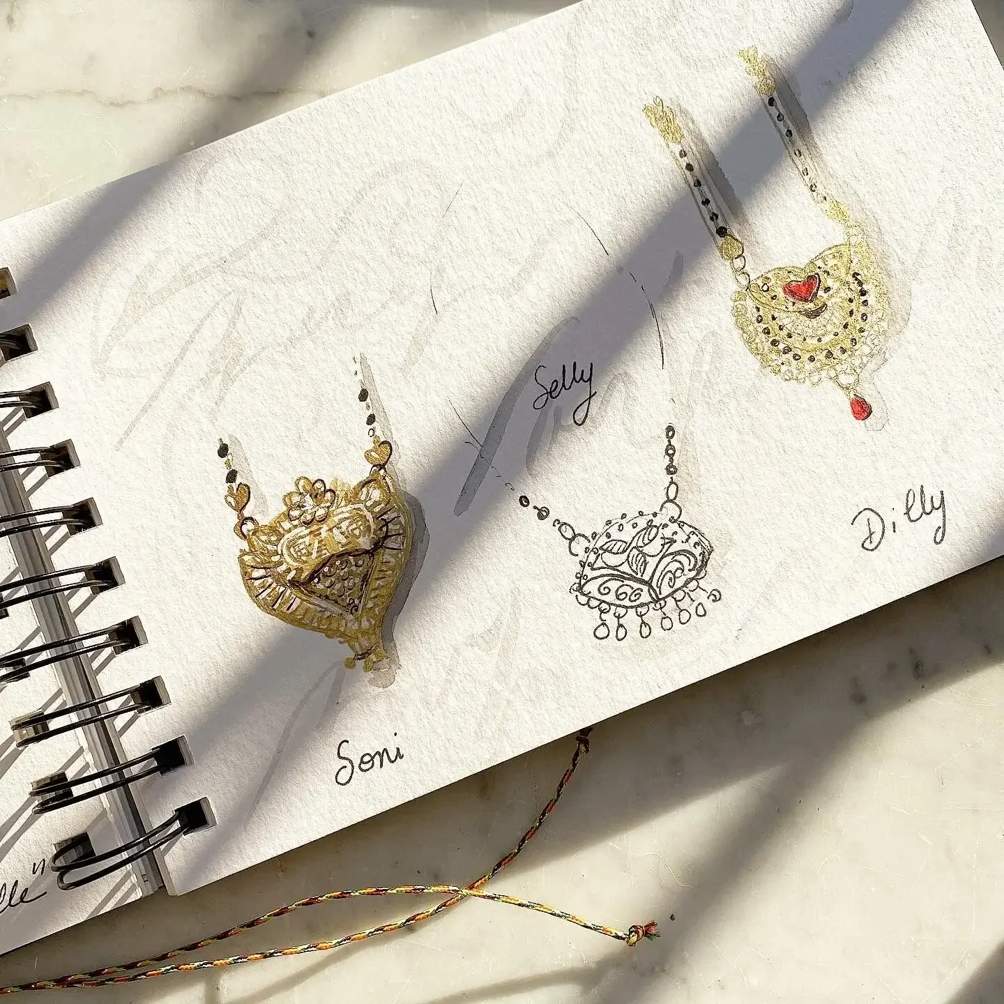 Dessins de colliers de bijoux avec des noms écrits dessous, Soni, Seily, Dily, sur une page d'un carnet. Ombres de branches et lumière naturelle.