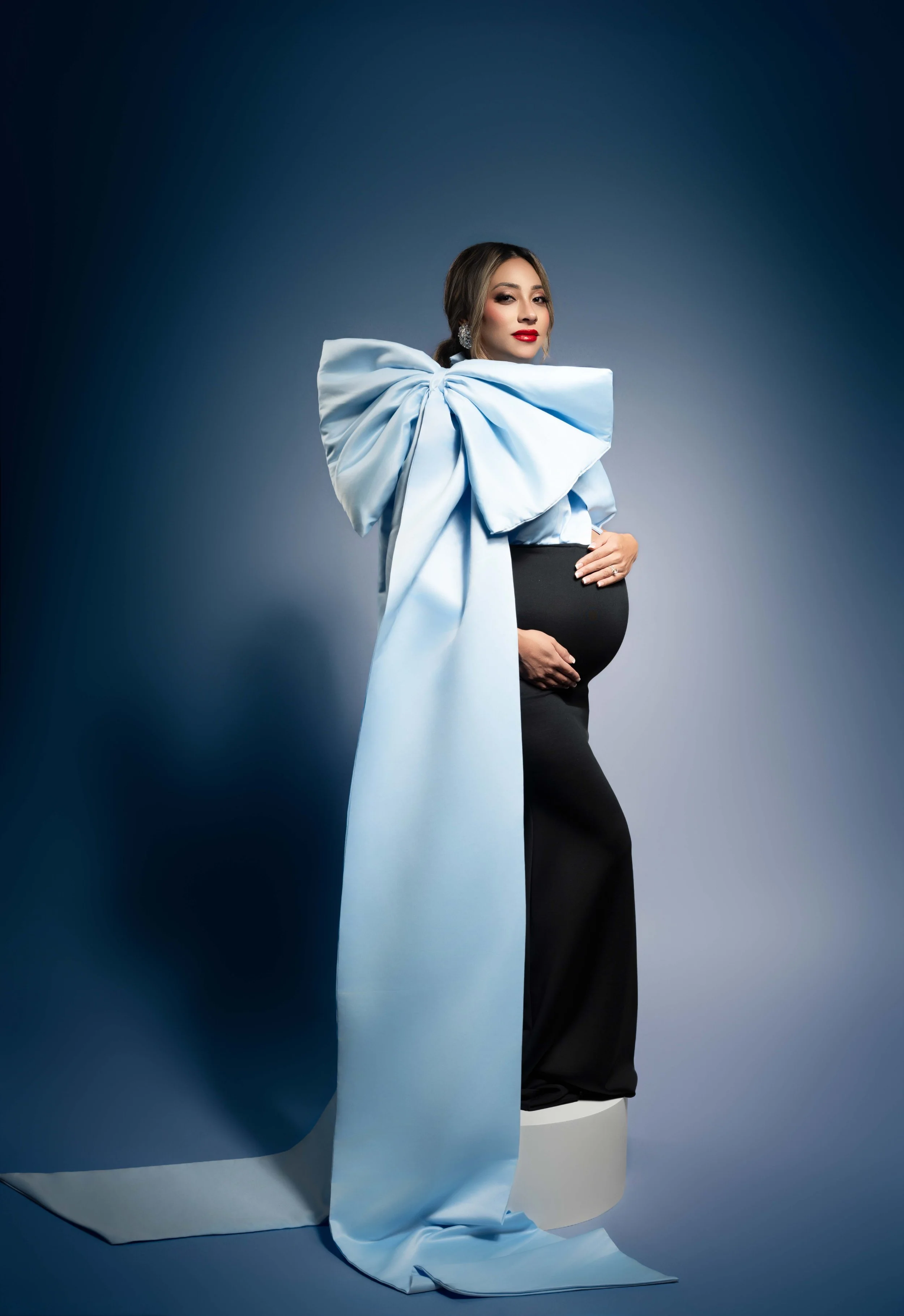 Pregnant woman in a black gown with an oversized pale blue bow trailing to the floor on a navy backdrop, fashion maternity portrait