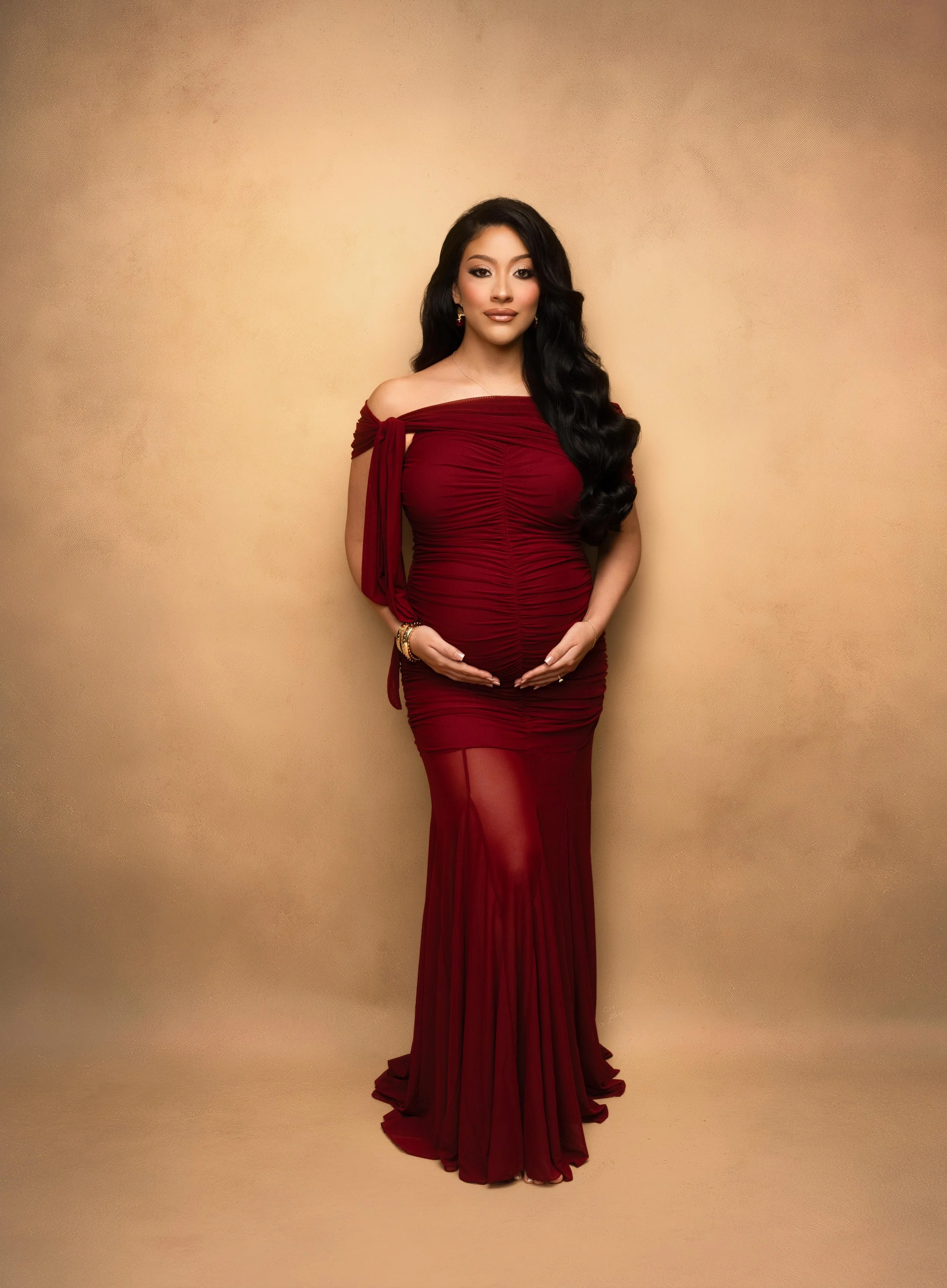 Pregnant woman in a red off-the-shoulder gown posing for an editorial maternity portrait against a warm neutral backdrop at Ana Lanier Portrait Studio in Dallas.