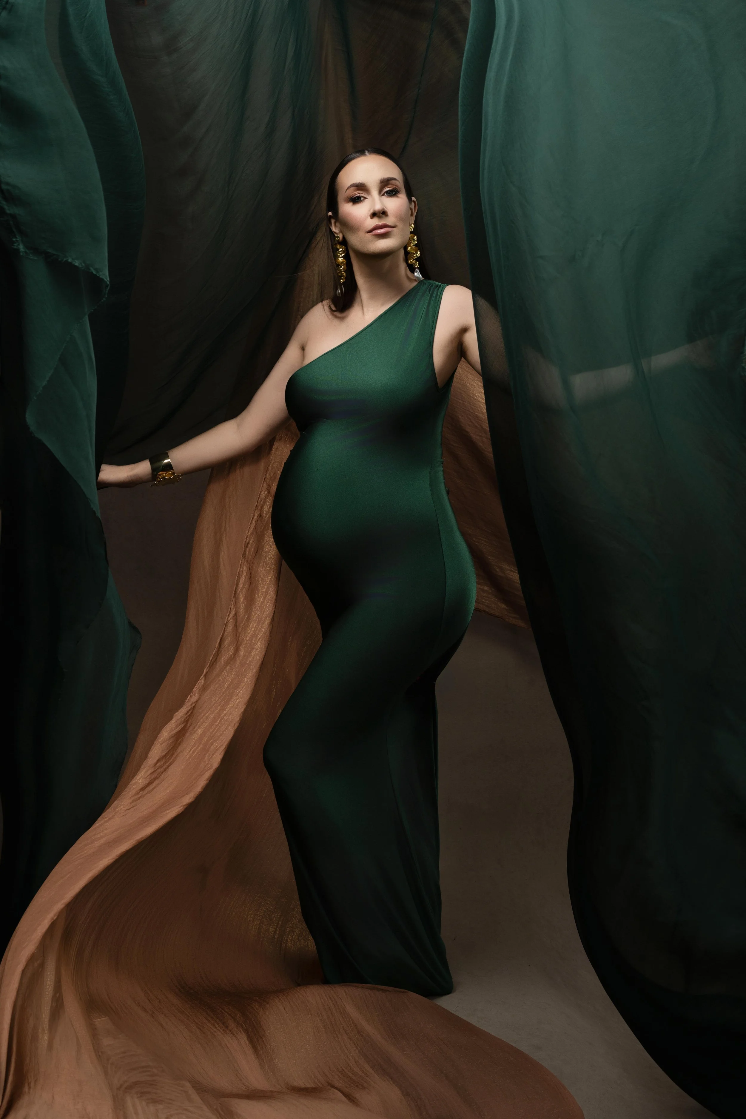 Fashion-inspired maternity portrait in an emerald green gown with flowing fabric, photographed in a Dallas studio.