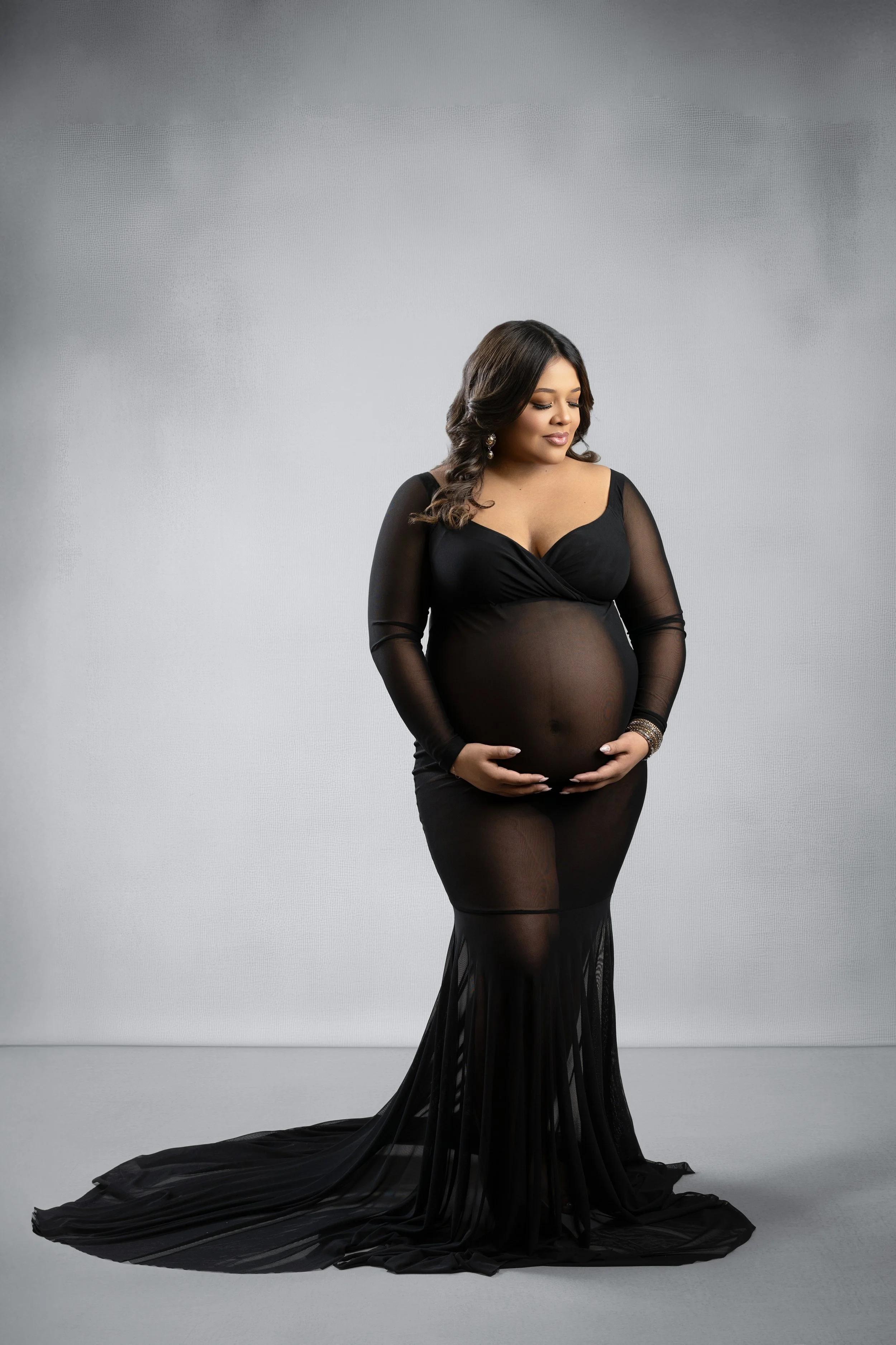 Pregnant woman in a sheer black long-sleeve gown with a train against a grey backdrop, maternity portrait