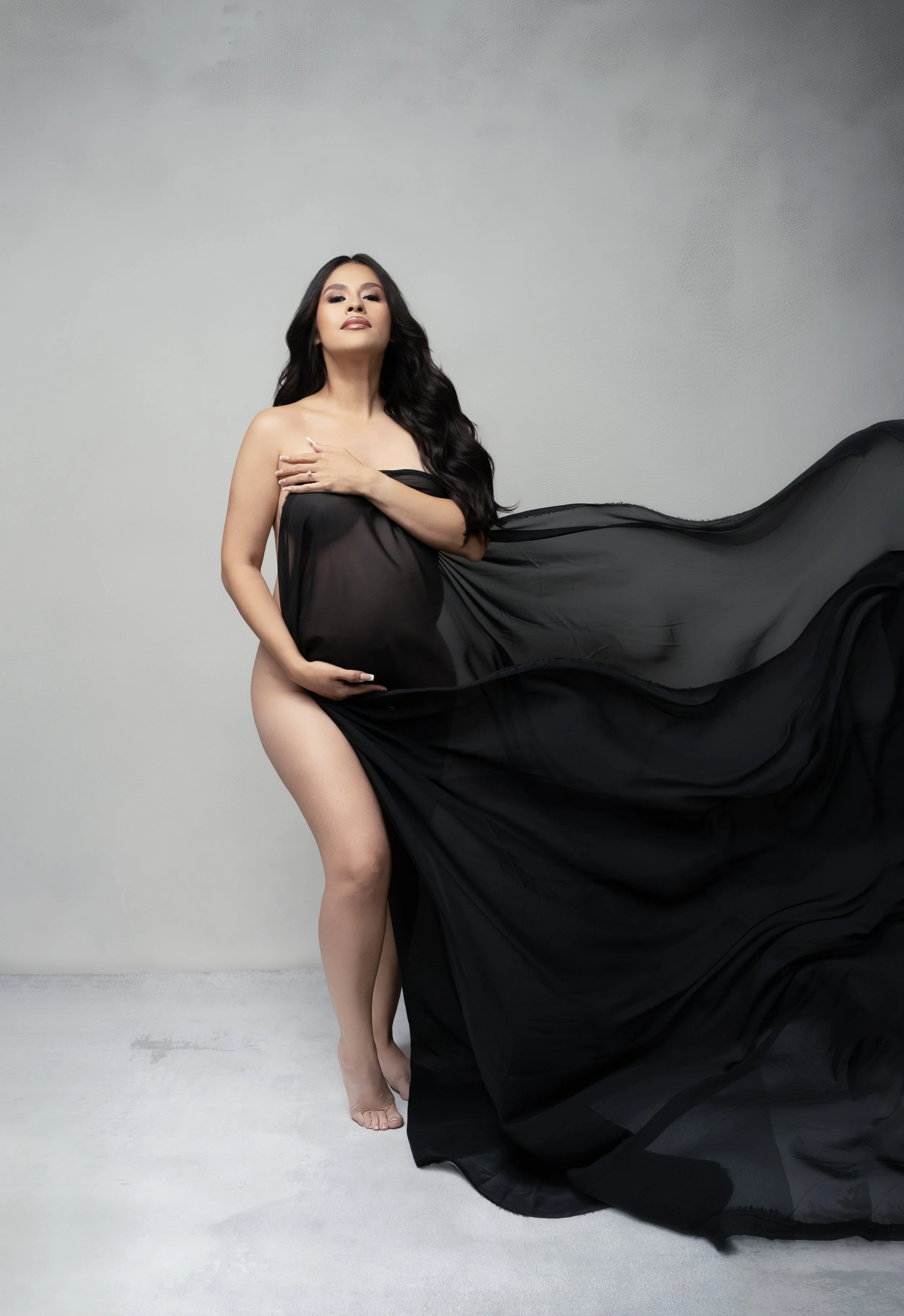 Pregnant woman holding sheer black fabric that sweeps out behind her, dramatic studio maternity portrait