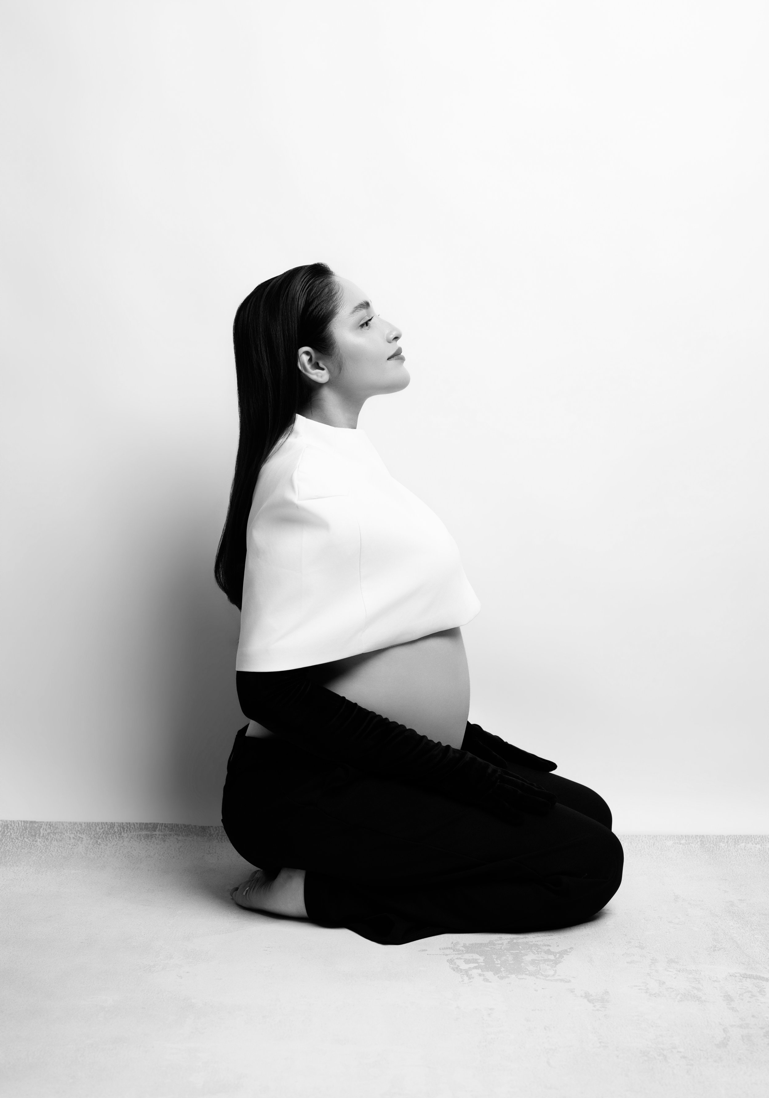 Black-and-white profile of a pregnant woman kneeling in a white cape top and long black gloves, minimalist studio maternity portrait