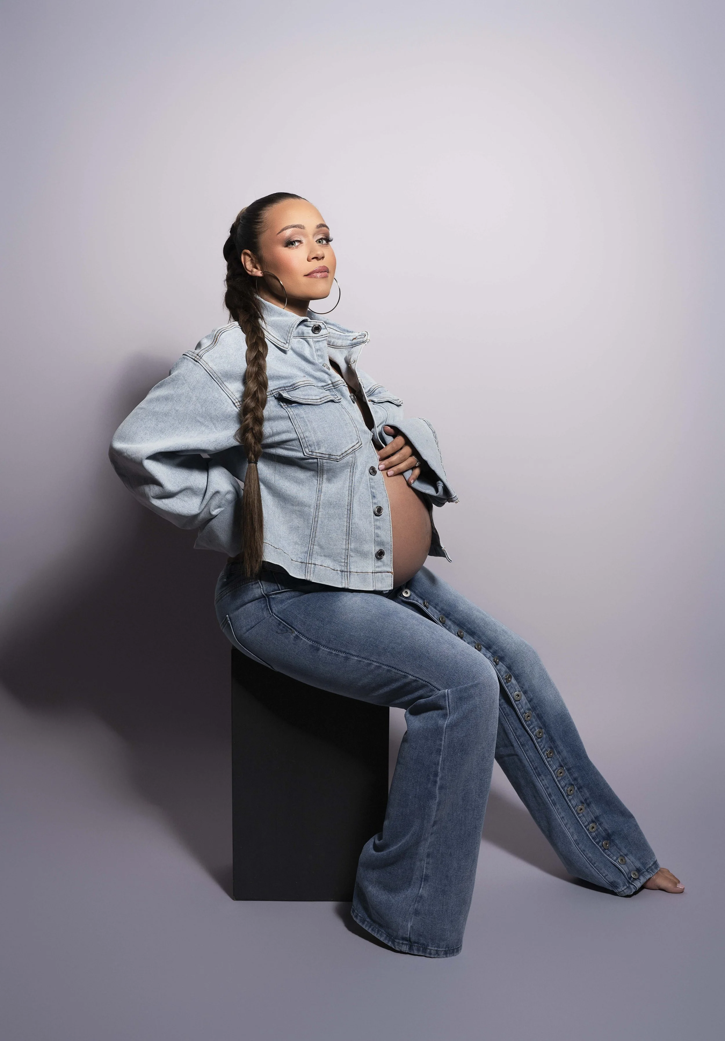 Pregnant woman with long braids in an oversized denim jacket and wide-leg jeans, seated, editorial maternity portrait