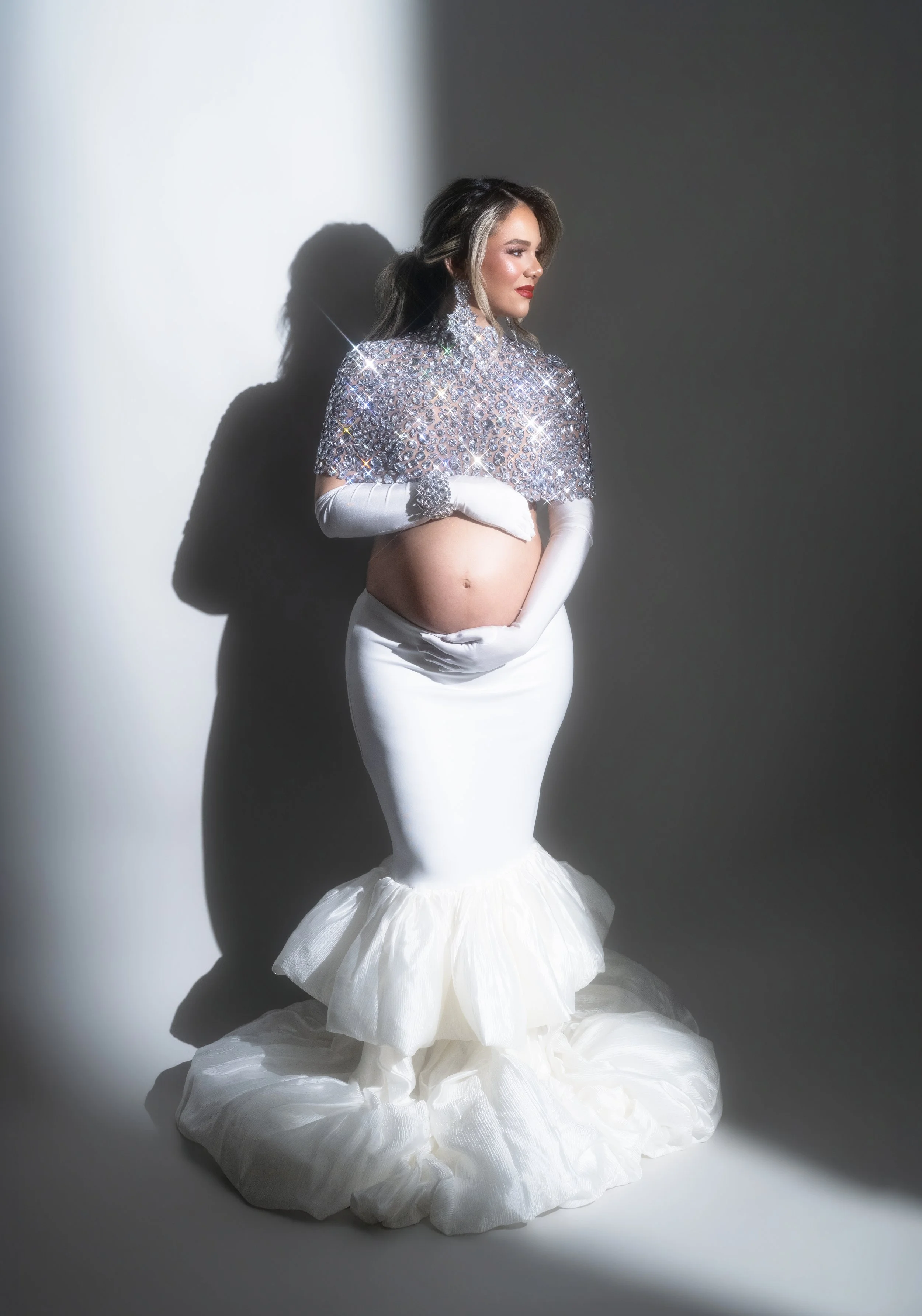 Pregnant woman in a white mermaid gown with a beaded silver top and white gloves, studio maternity portrait with hard shadow