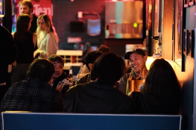 A group of people sitting at a table in a dimly lit restaurant or bar, with some standing in the background, engaged in conversation and smiling.