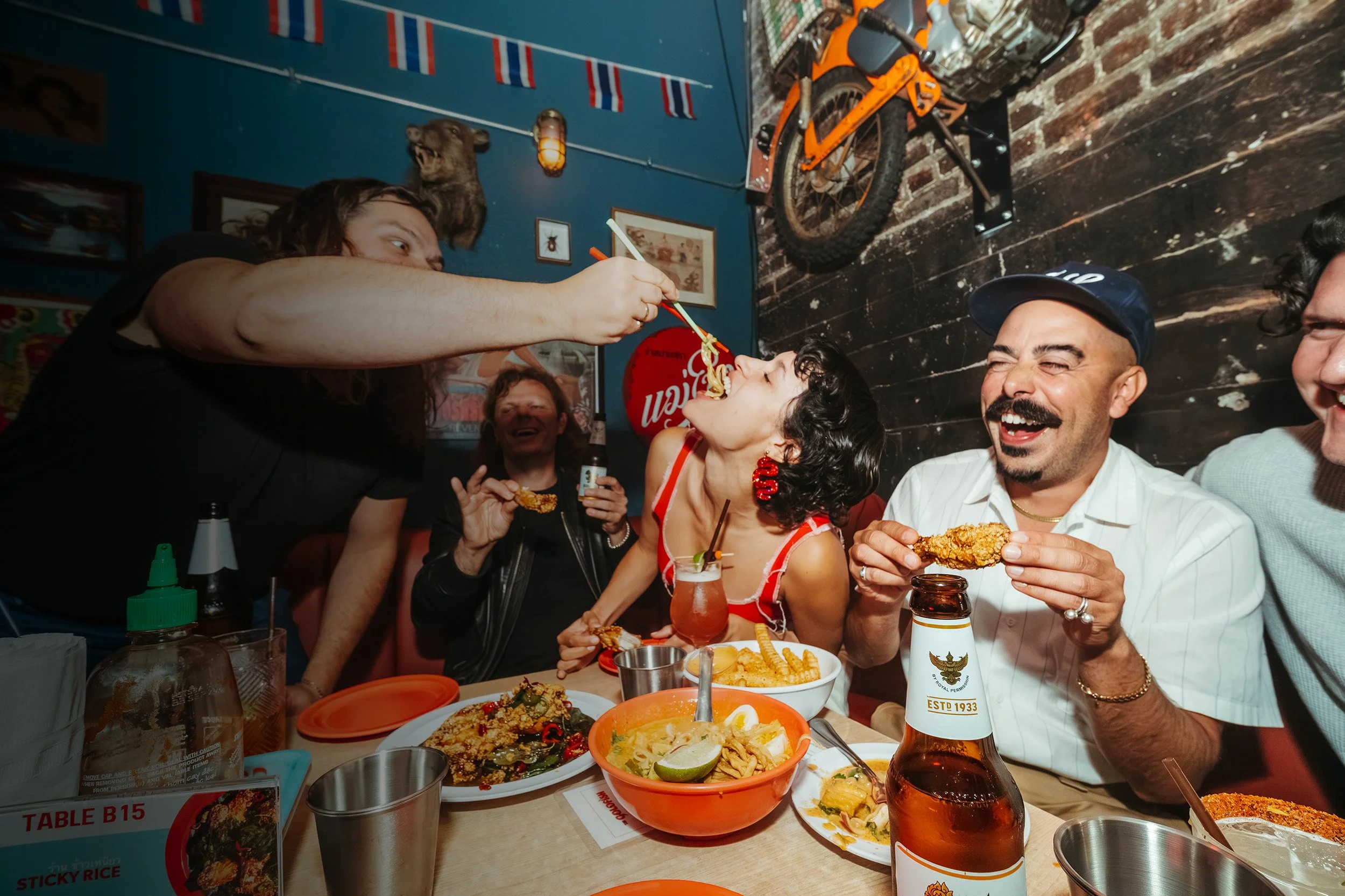 People enjoying food at a restaurant, with one person feeding noodles to another, who is opening their mouth for the food; food on the table includes fried chicken, noodles, and drinks; the setting has Asian decor.