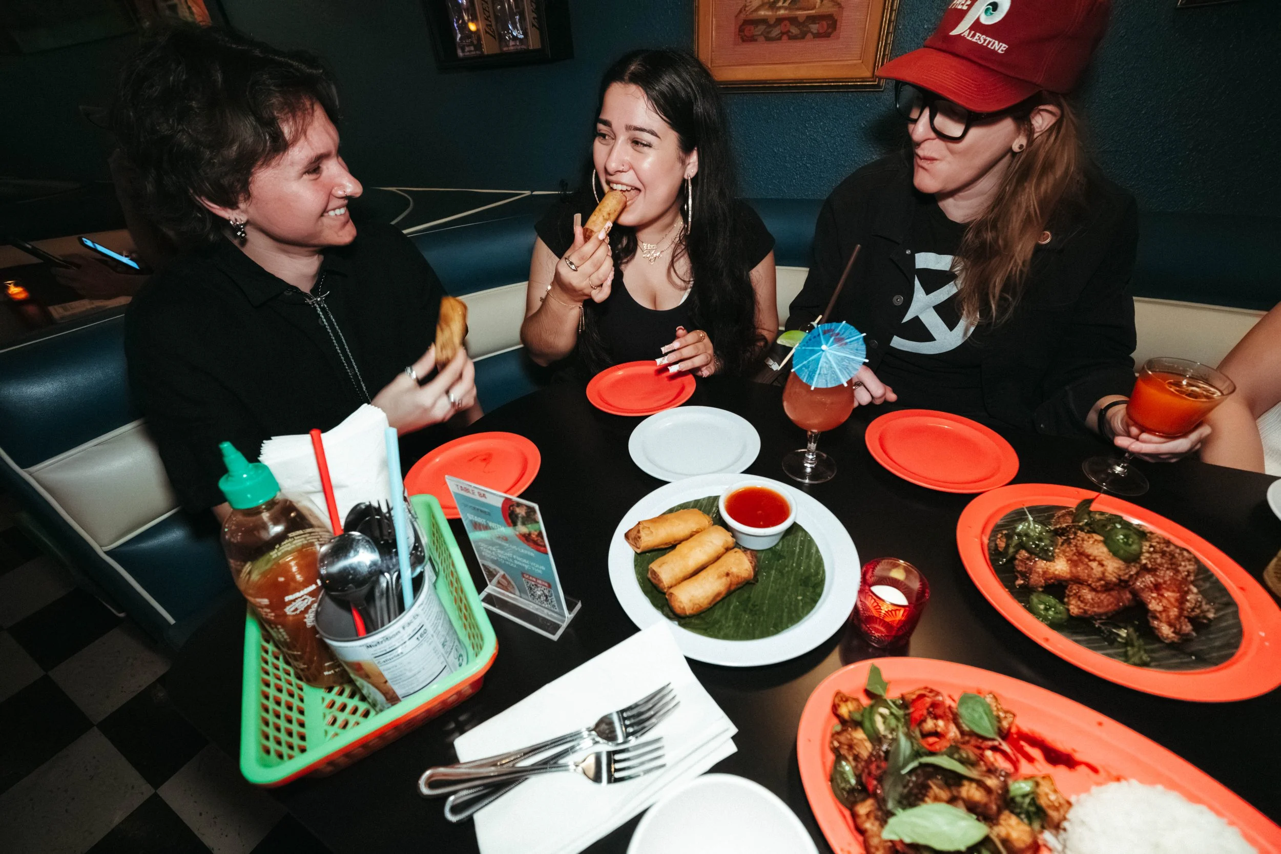 Three women sitting at a restaurant table enjoying Asian food and drinks, with sushi rolls, spring rolls, fried chicken, rice, and various beverages including cocktails and water.