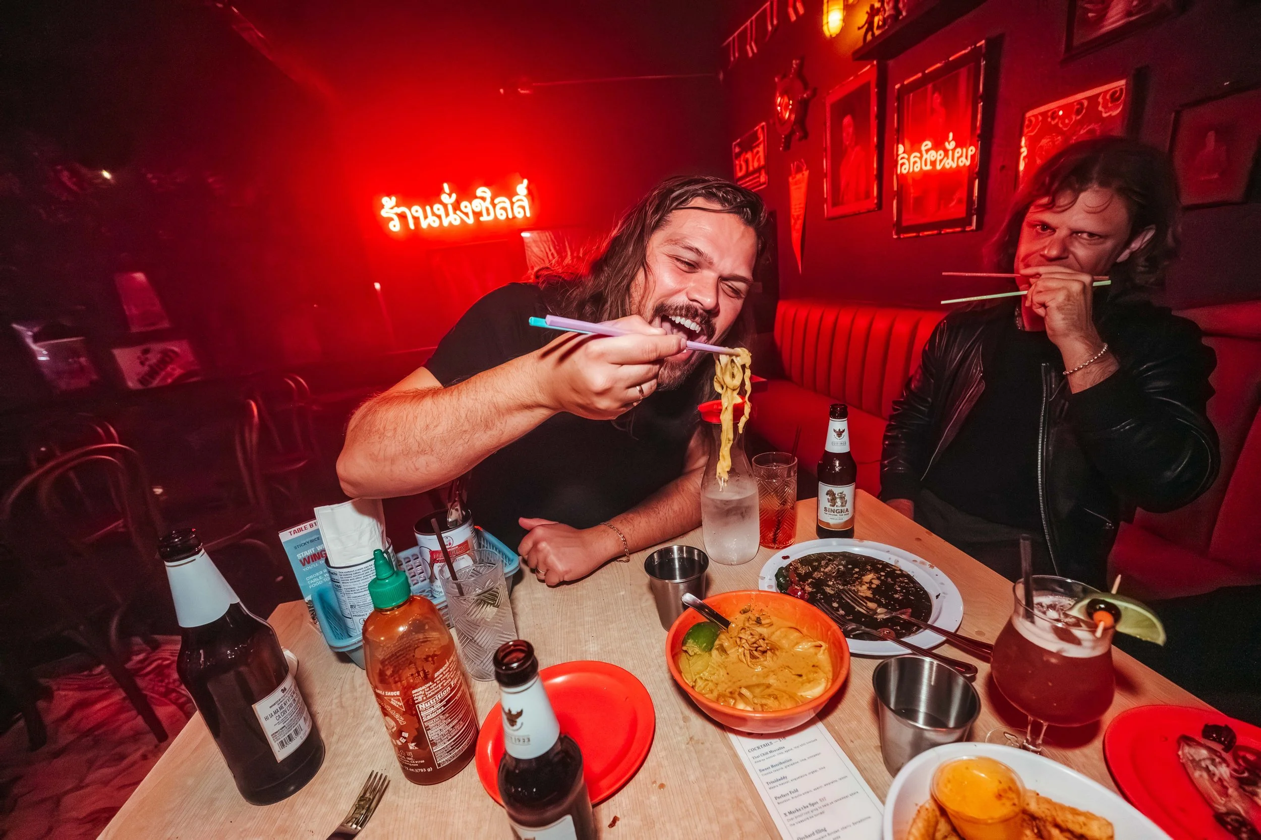 Two men dining at a restaurant, one is eating noodles with chopsticks and the other is holding chopsticks near his mouth, surrounded by various dishes and drinks with red neon signs on the wall behind them.