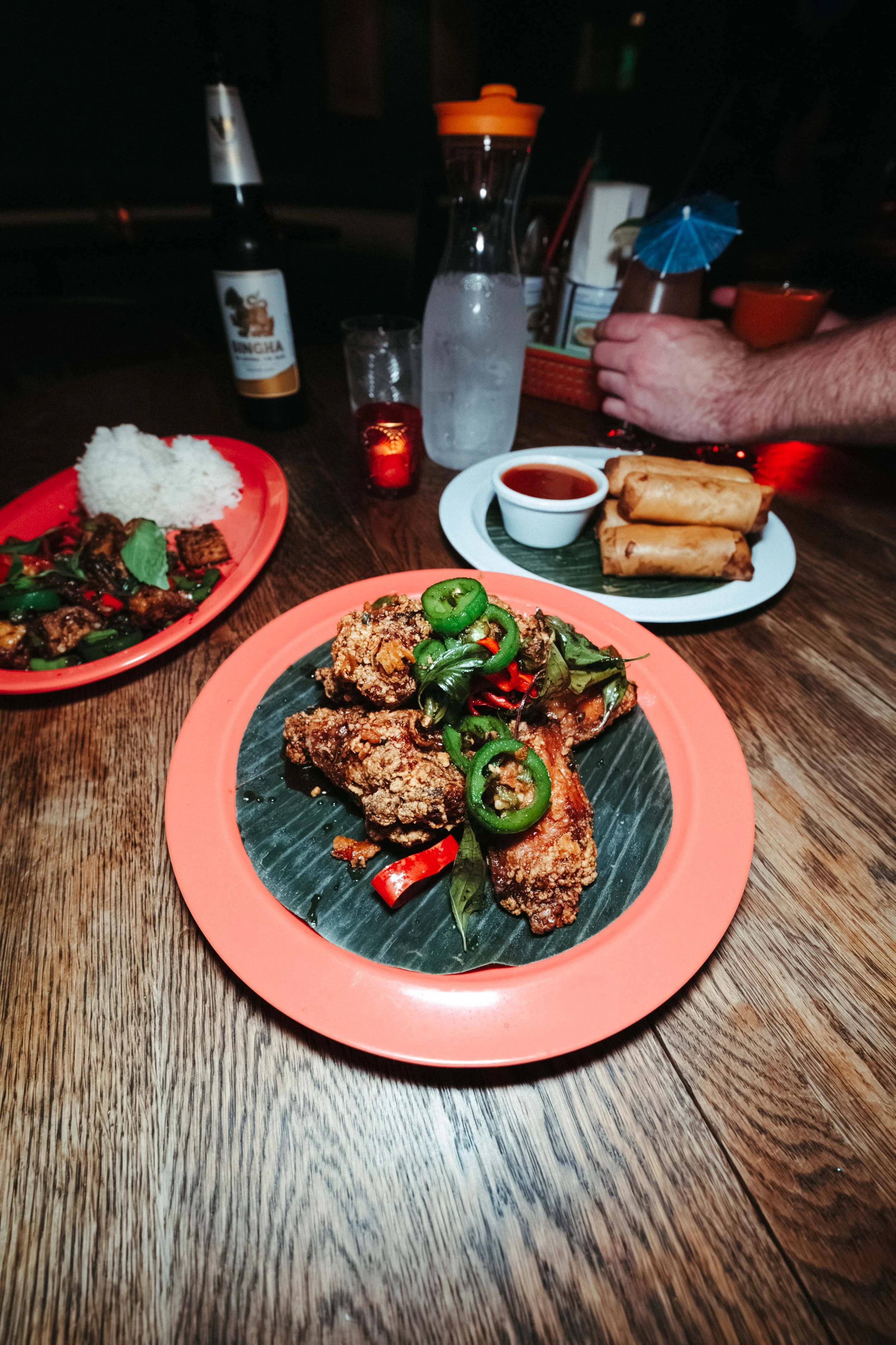 Fried chicken topped with sliced green peppers and red chili on a pink plate, with rice and stir-fried vegetables in the background, on a wooden table.