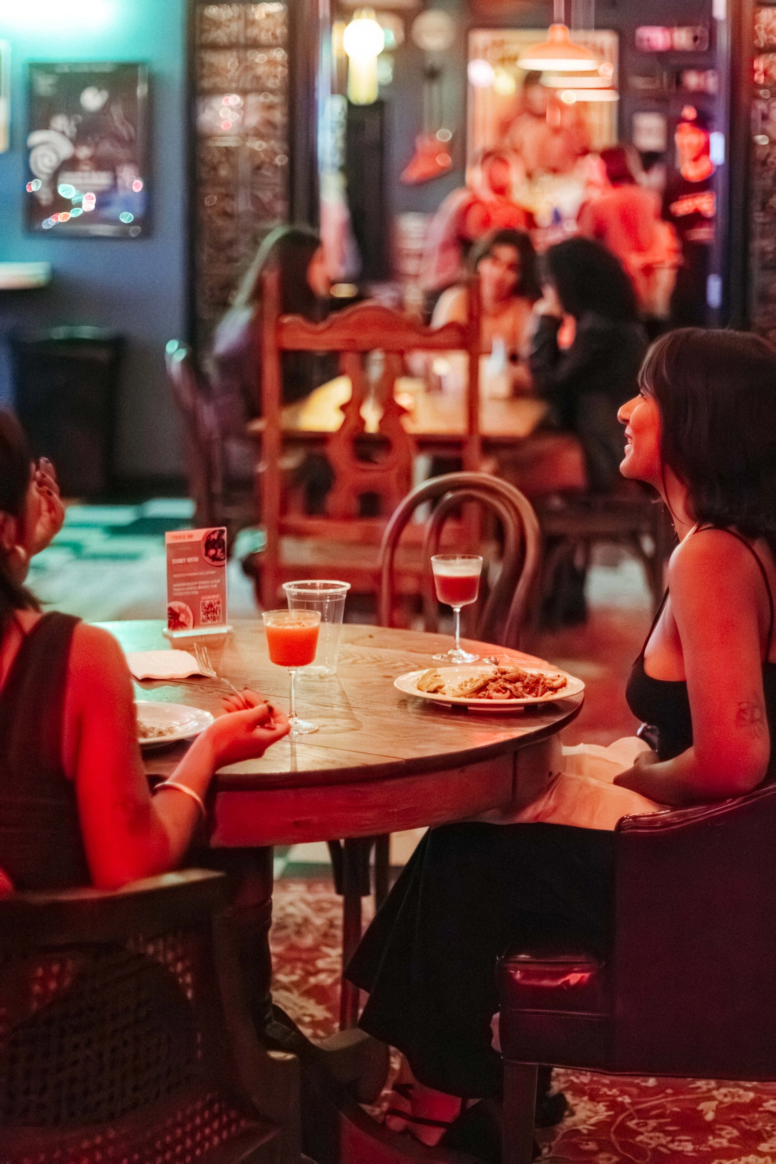 Two women sitting at a wooden table in a dimly lit restaurant or bar, enjoying drinks and pasta, with other patrons in the background.
