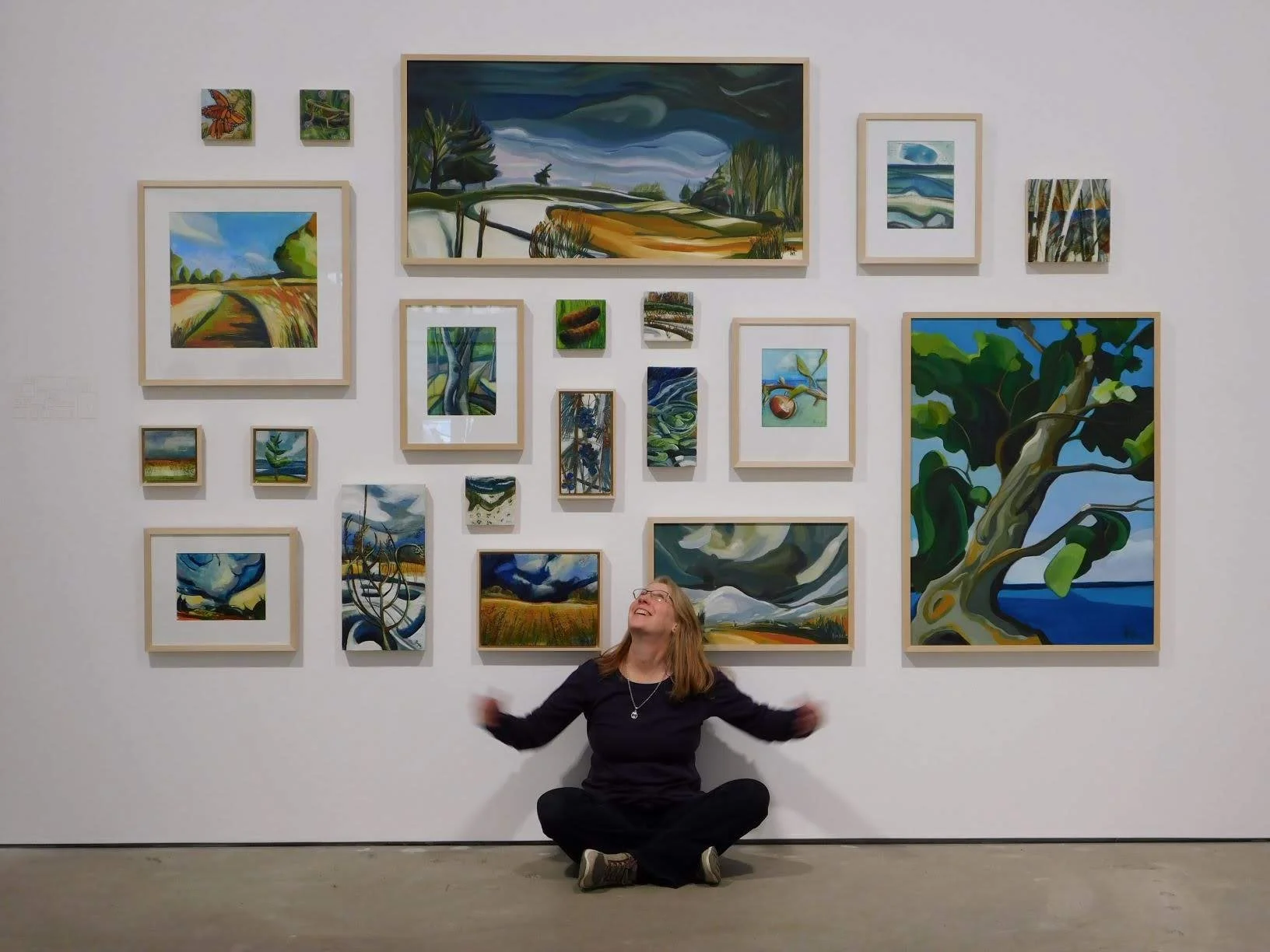 A woman sitting on the floor in front of a white wall with multiple colorful landscape paintings, smiling and looking up.