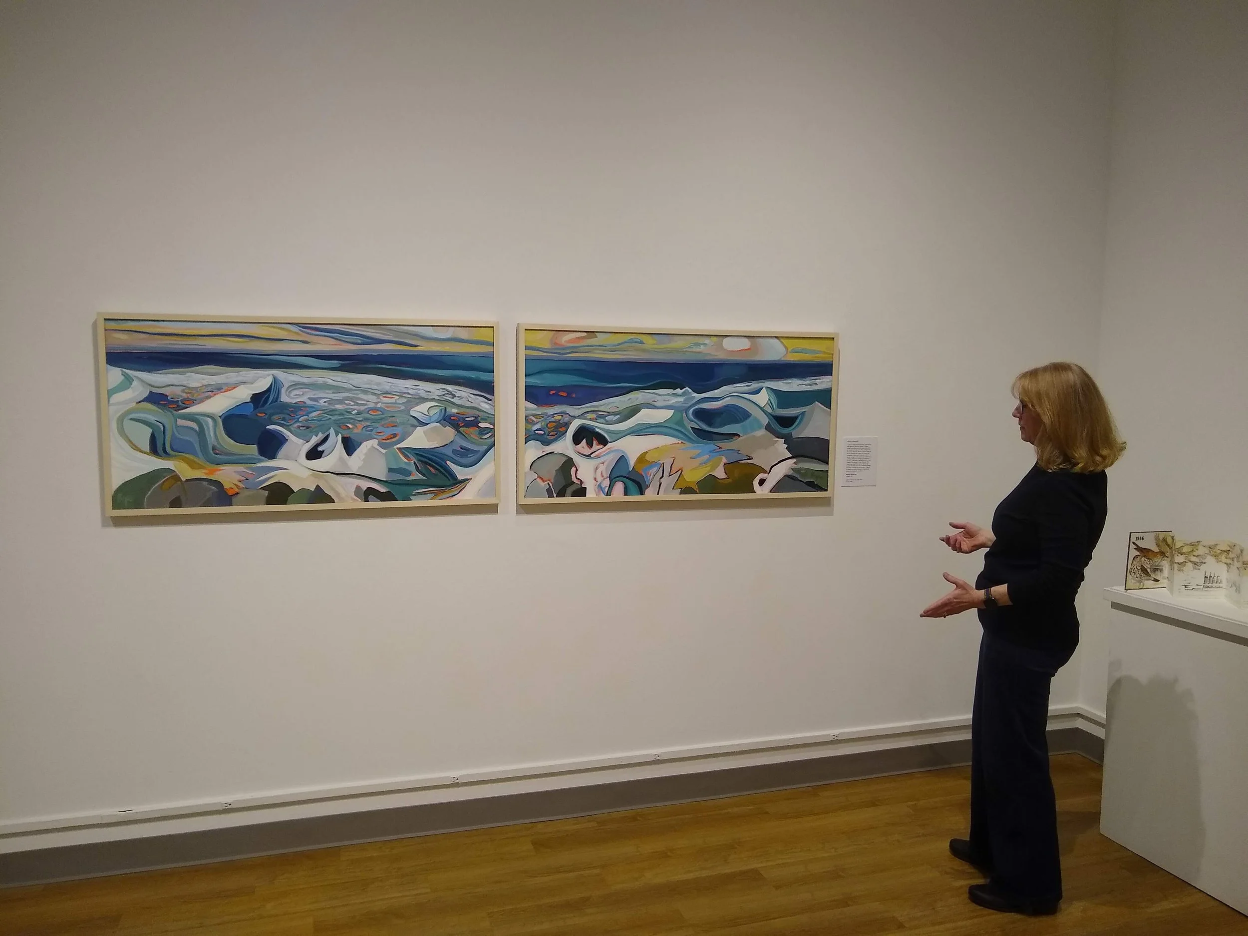 A woman with blonde hair wearing glasses, a black top, and black pants standing in an art gallery, looking at two abstract landscape paintings with swirling blues, whites, and other colors on the wall. There is a small white table with open books or pamphlets to her right.