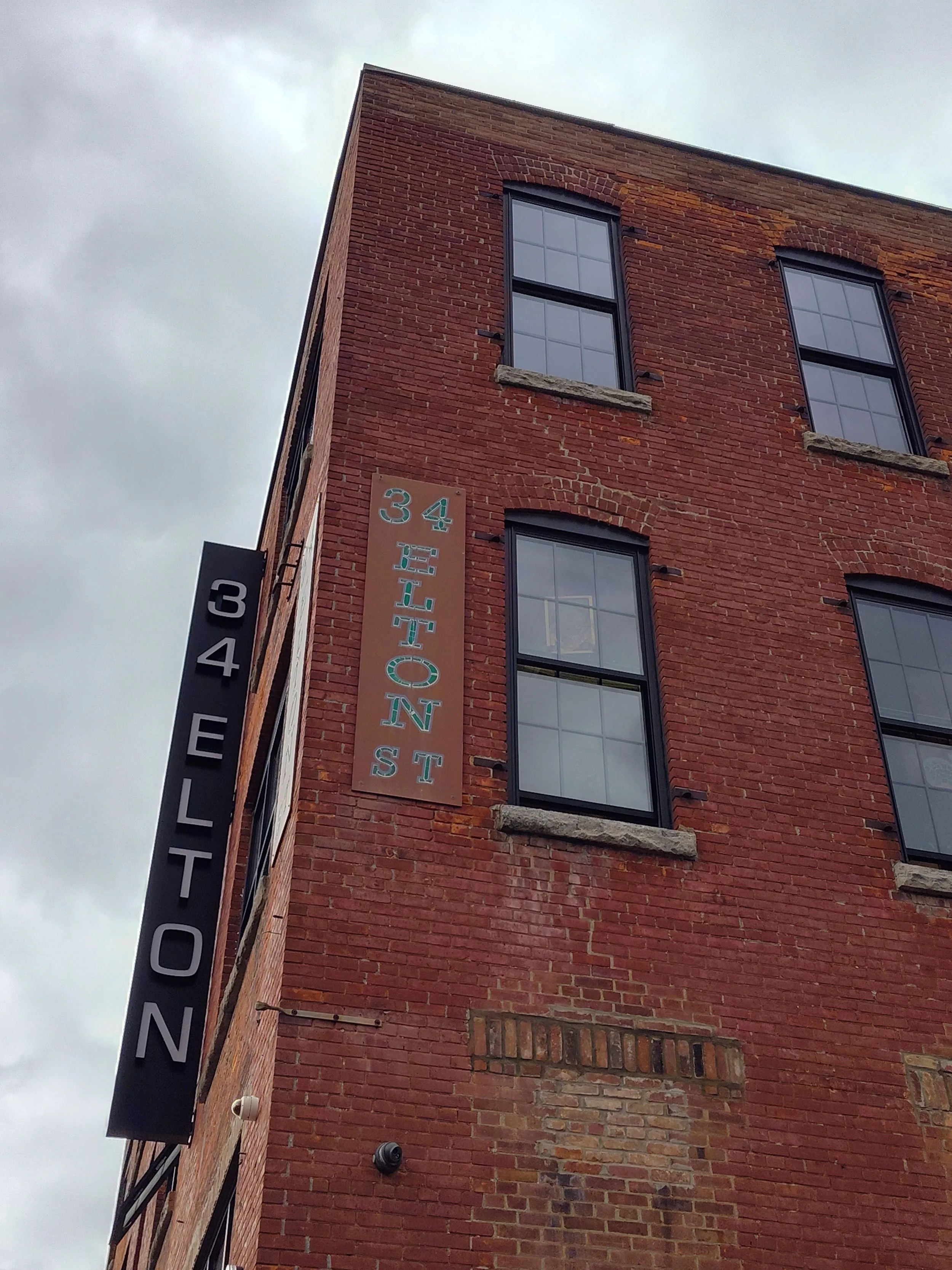 Brick building with black window frames and two signs, one vertical with the text '34ELTON' and another horizontal with the text '34TH ST'.