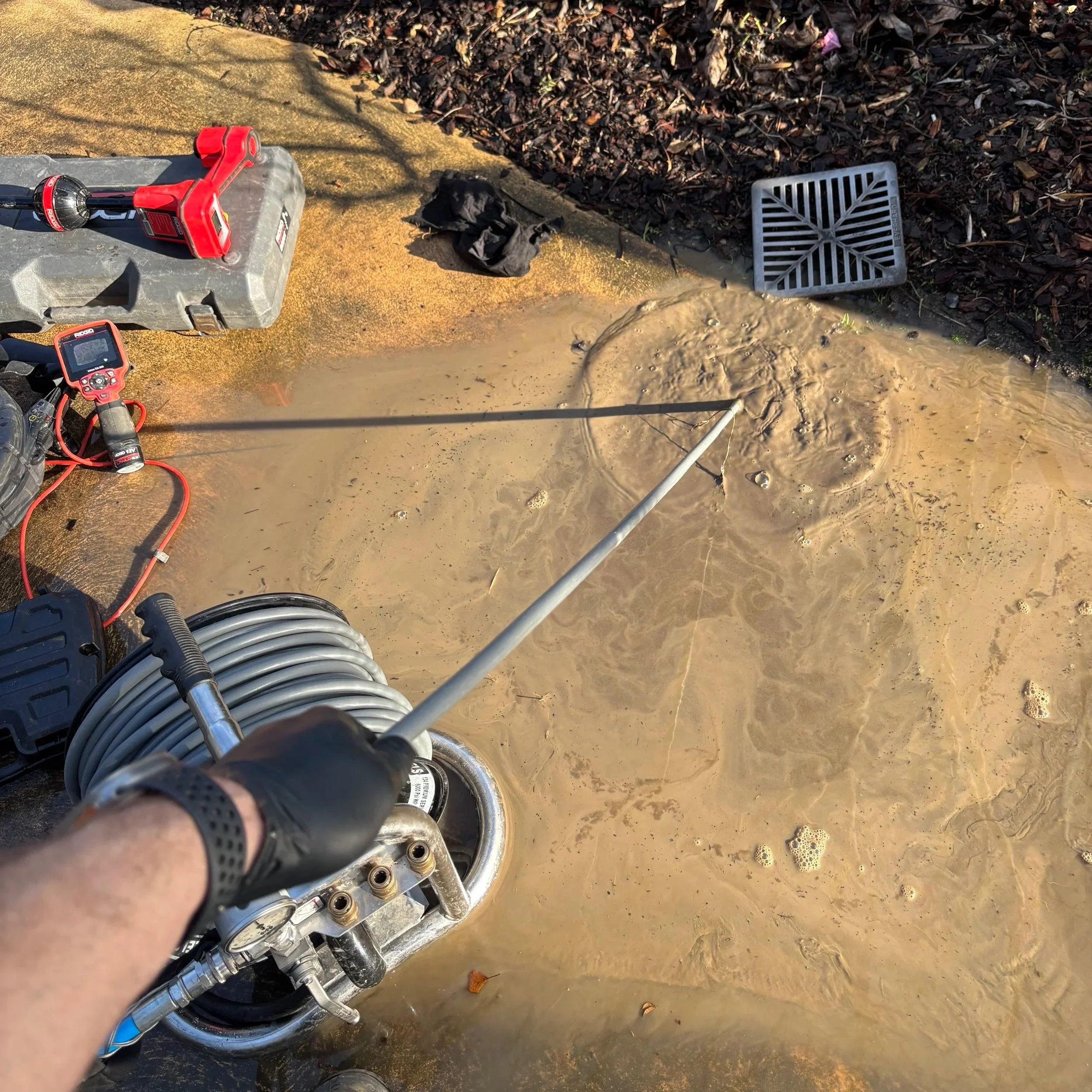 Photo of a person riding an off-road vehicle through a muddy area, with focus on the vehicle's metal wheel and various tools and equipment attached, including a red cordless drill and a tool case, near a drain grate on the muddy ground.