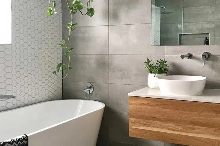 Modern bathroom with gray tiled walls, a white freestanding bathtub, and a wooden vanity with a vessel sink and a small green plant.