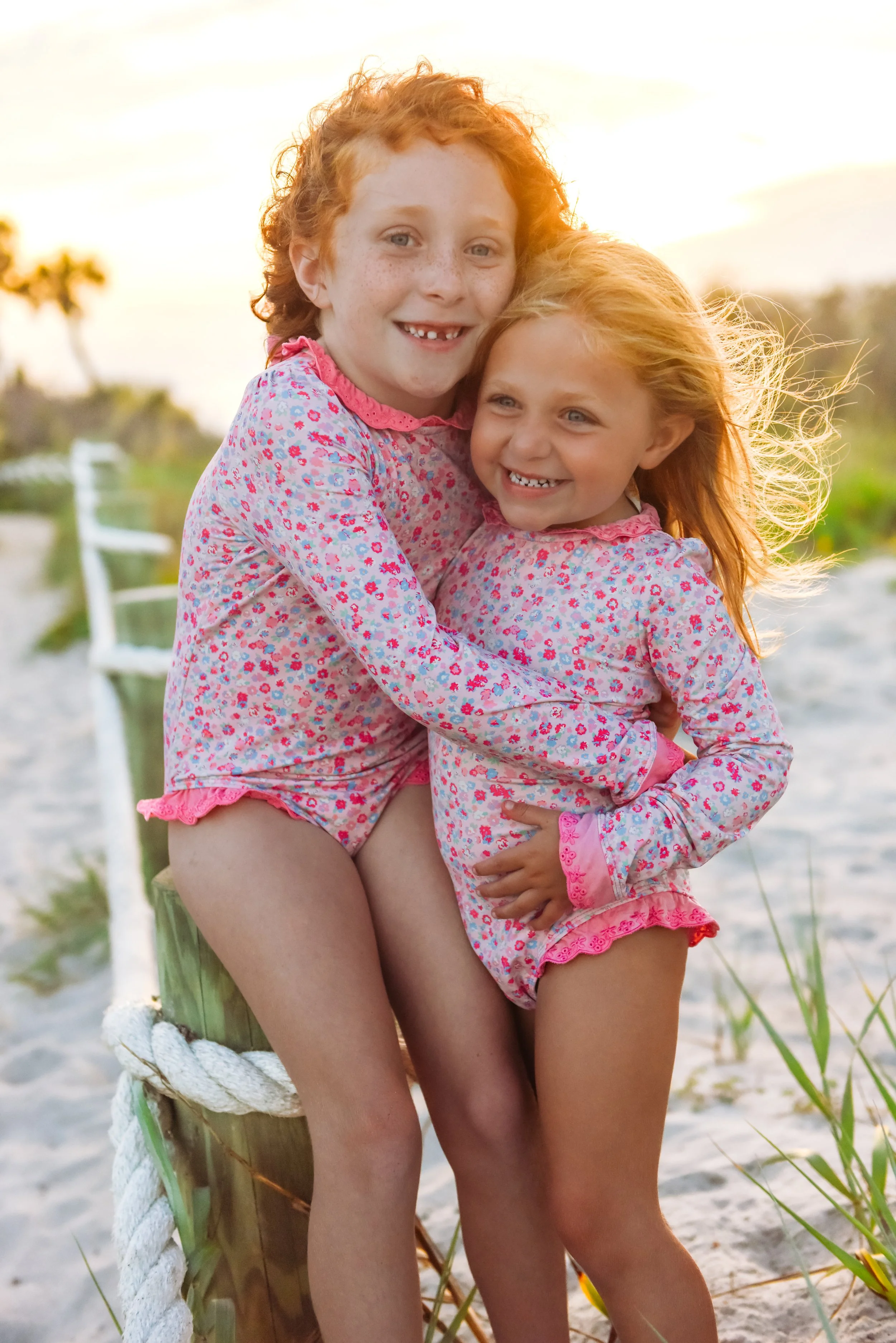 Two young girls with red hair, wearing matching floral pajamas, sitting on a weathered wooden post at the beach. The older girl is hugging the younger girl, both smiling happily, with the sunset behind them and sandy beach surroundings.