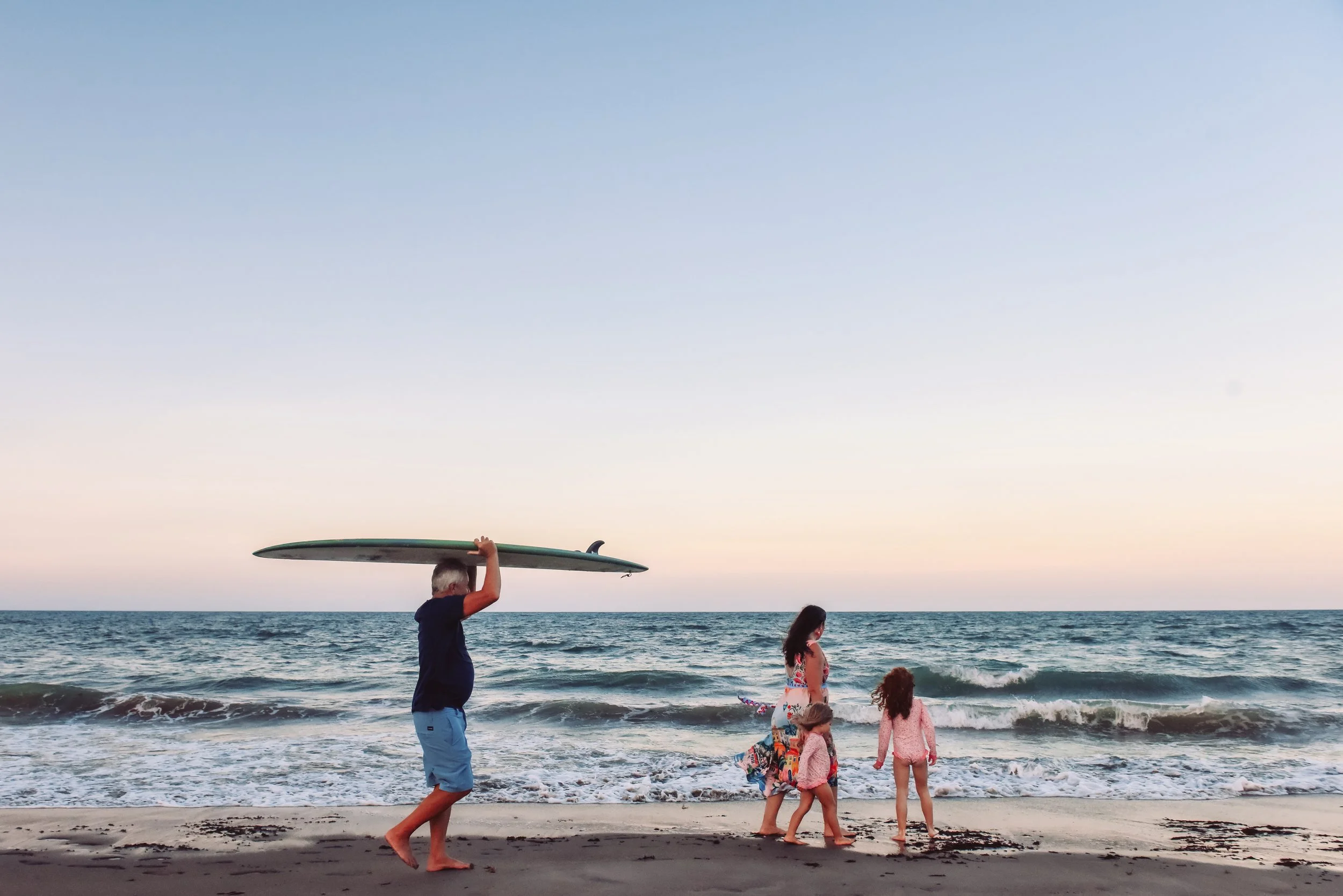 A man holding a surfboard walking along the beach at sunset, with children playing near the shoreline and the ocean waves in the background.