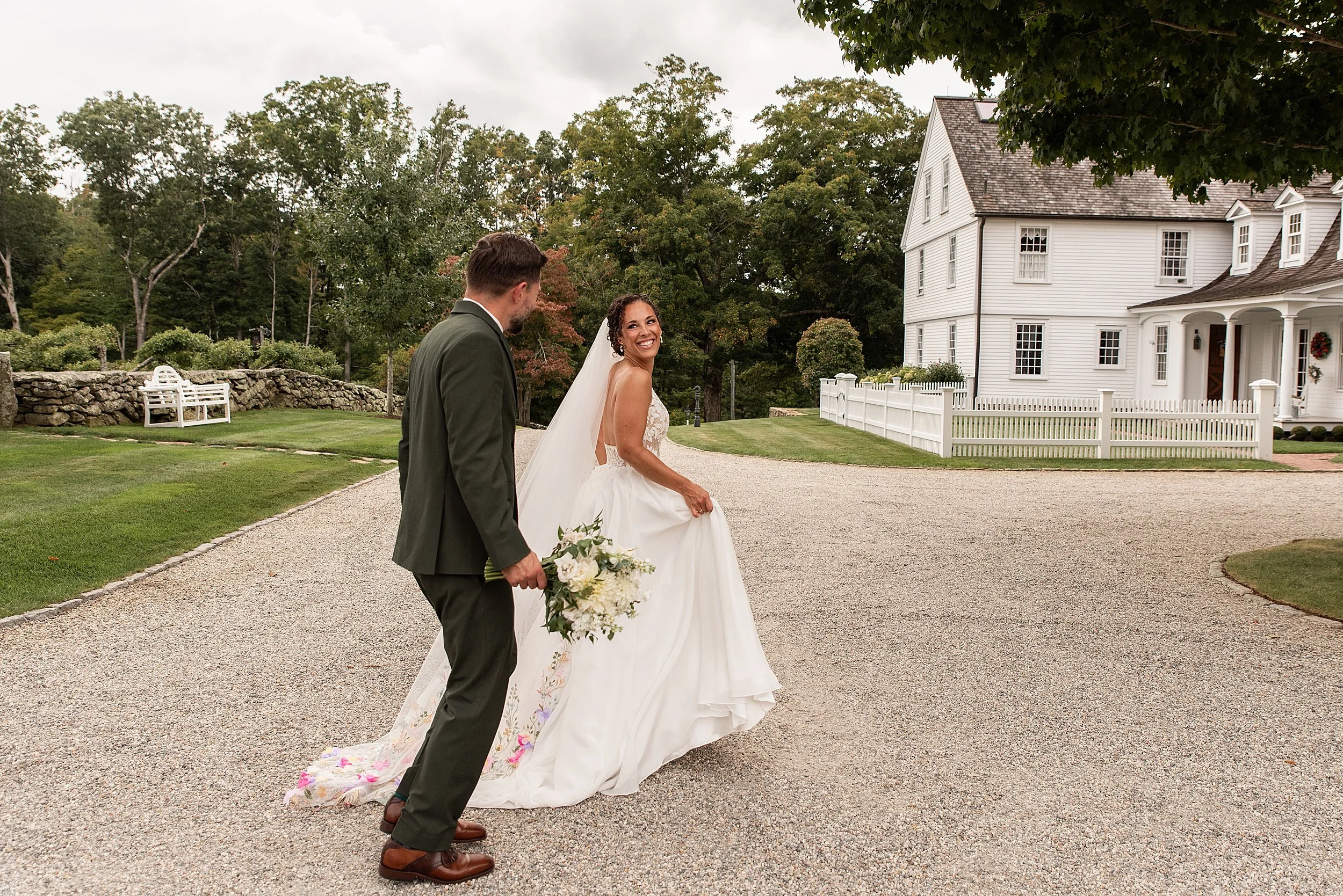 A bride holding her dress running in front of the historic cottage at Smith Farm Gardens as the groom holds her bouquet and runs beside her.