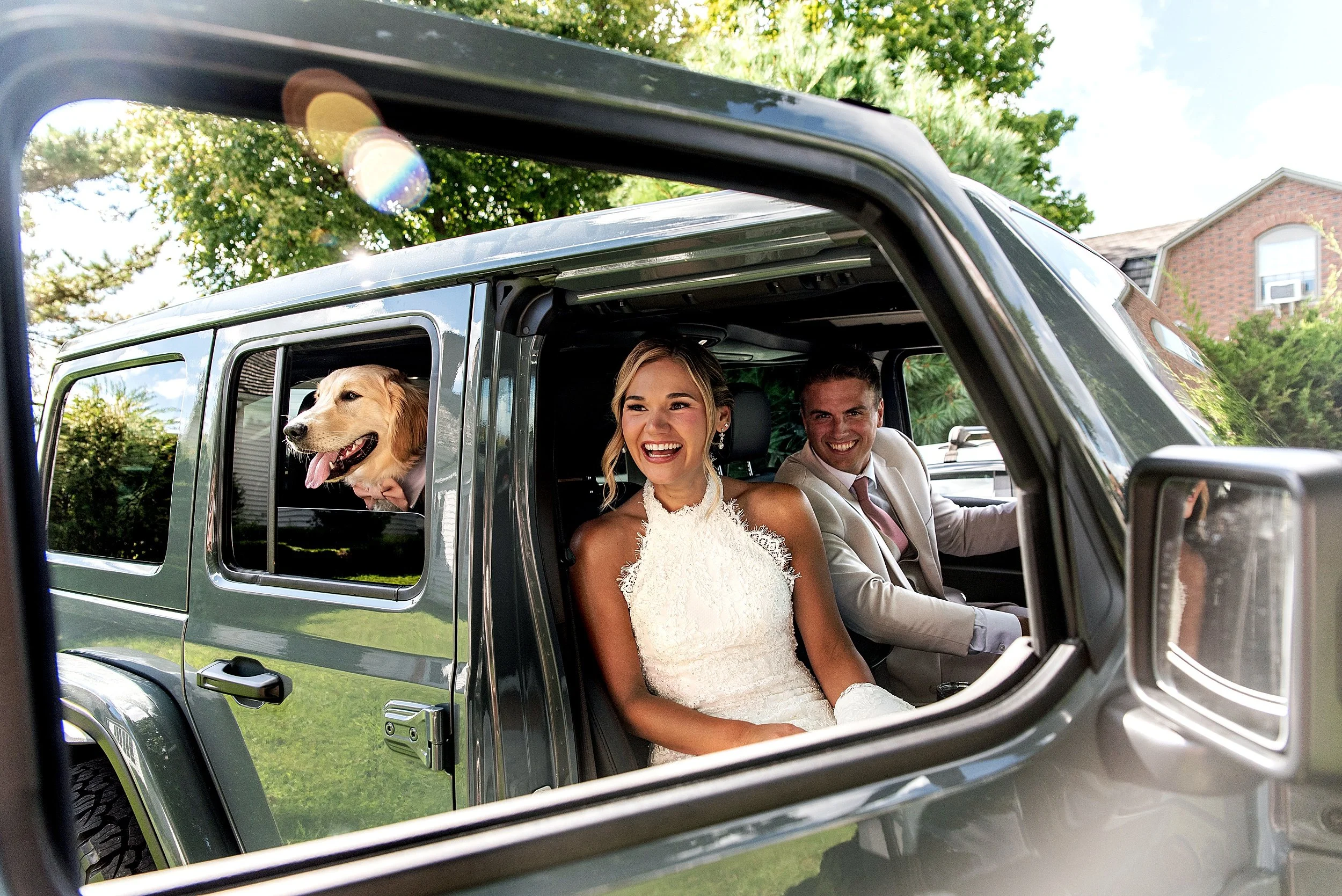 A fun photo captured through a vehicle window of a wedding couple in their Jeep with their dog sticking his head out.