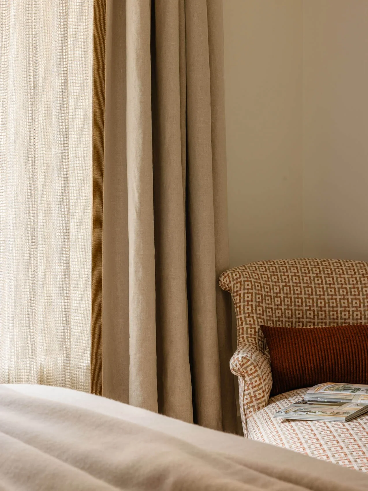 Bedroom detail from Paris apartment renovation showing layered sheer and fabric curtains, patterned accent chair, soft neutral textiles, and warm interior styling.