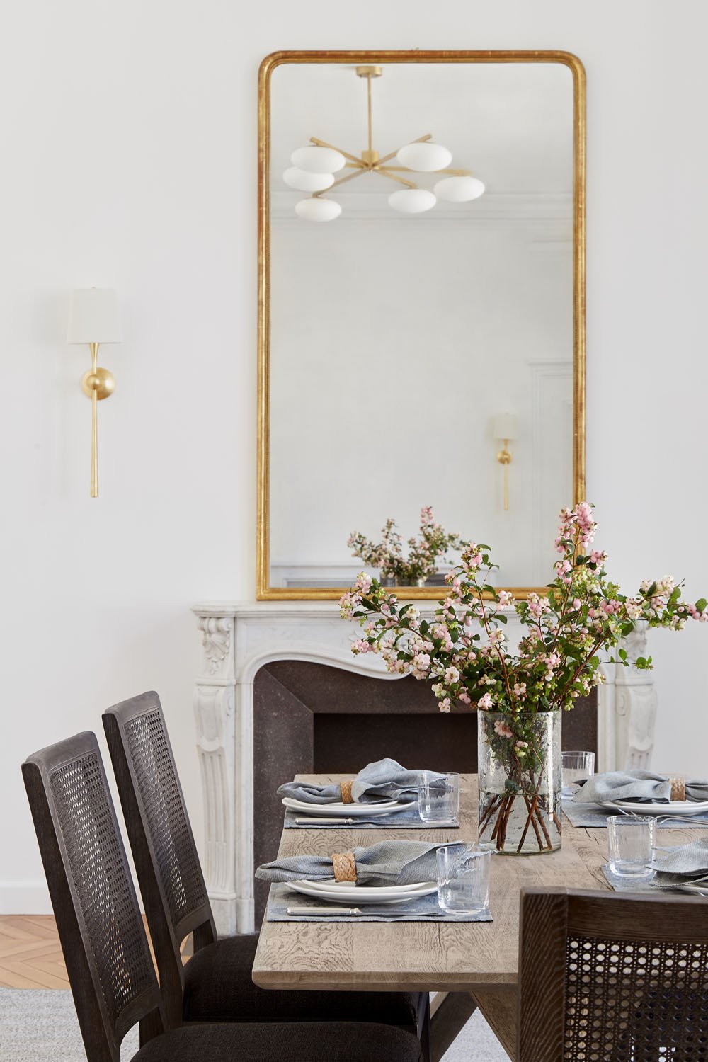  Parisian dining room with plates set up on the wooden table together with a beautiful flower arrangement. The white wall is decorated with a big mirror with golden frame. 