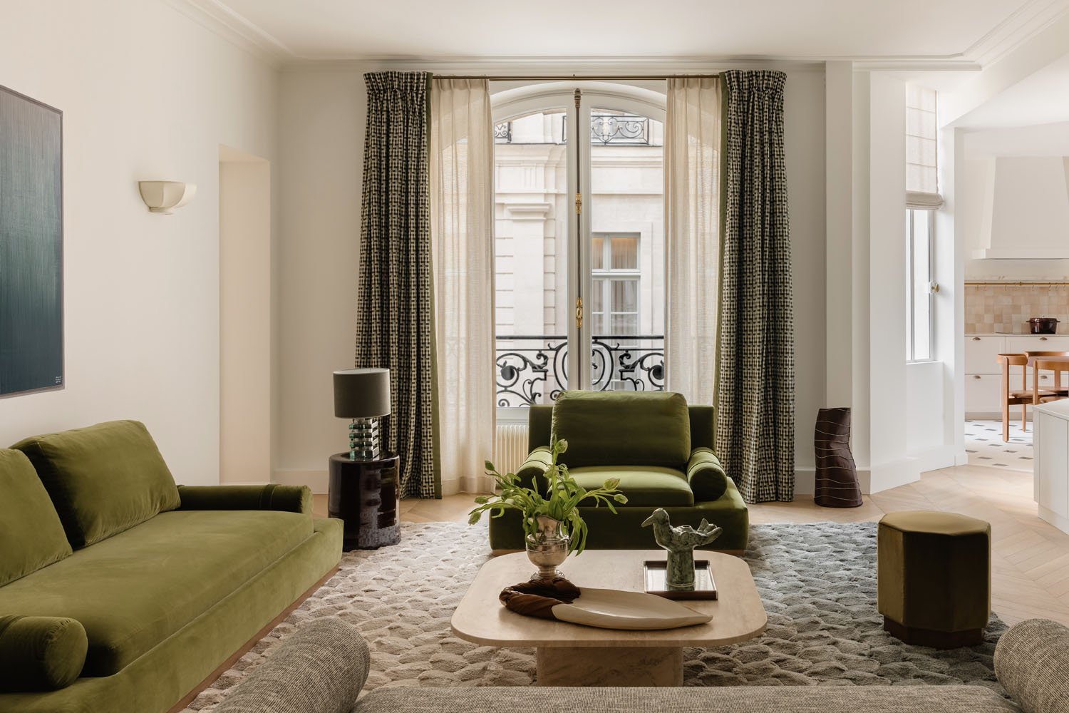 Modern Paris apartment living room renovation with green velvet seating, large French windows, neutral decor, and open-plan kitchen view on Rue de Lille.