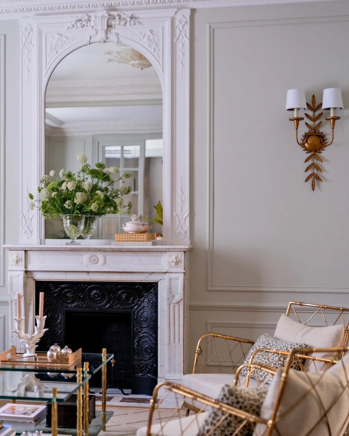 Parisian living room with ornate white marble fireplace, carved trumeau mirror, fresh floral arrangement, gold wall sconce and bamboo armchair