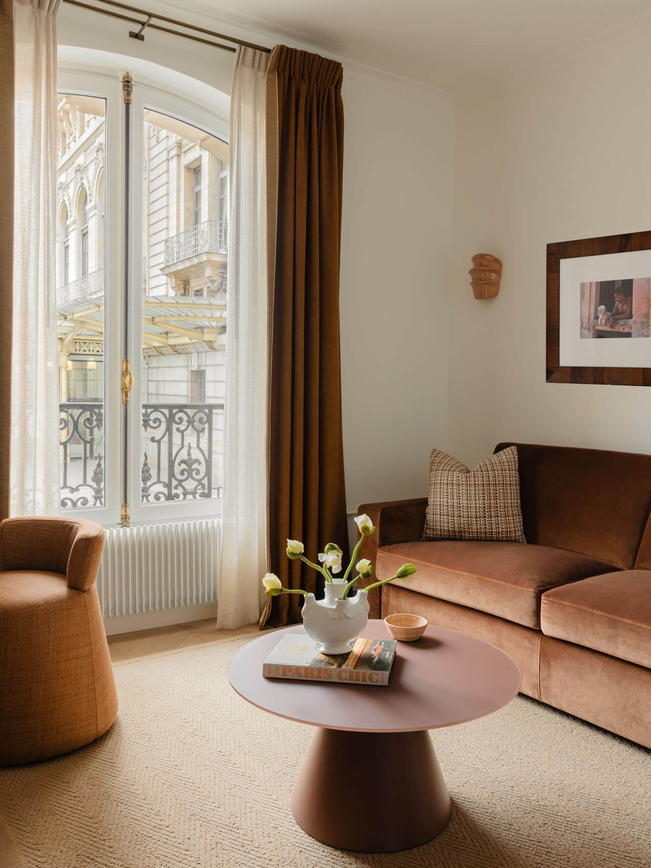 Paris apartment family room renovation with warm brown sofa, round modern coffee table, arched French window, sheer curtains, and classic wrought-iron balcony view.